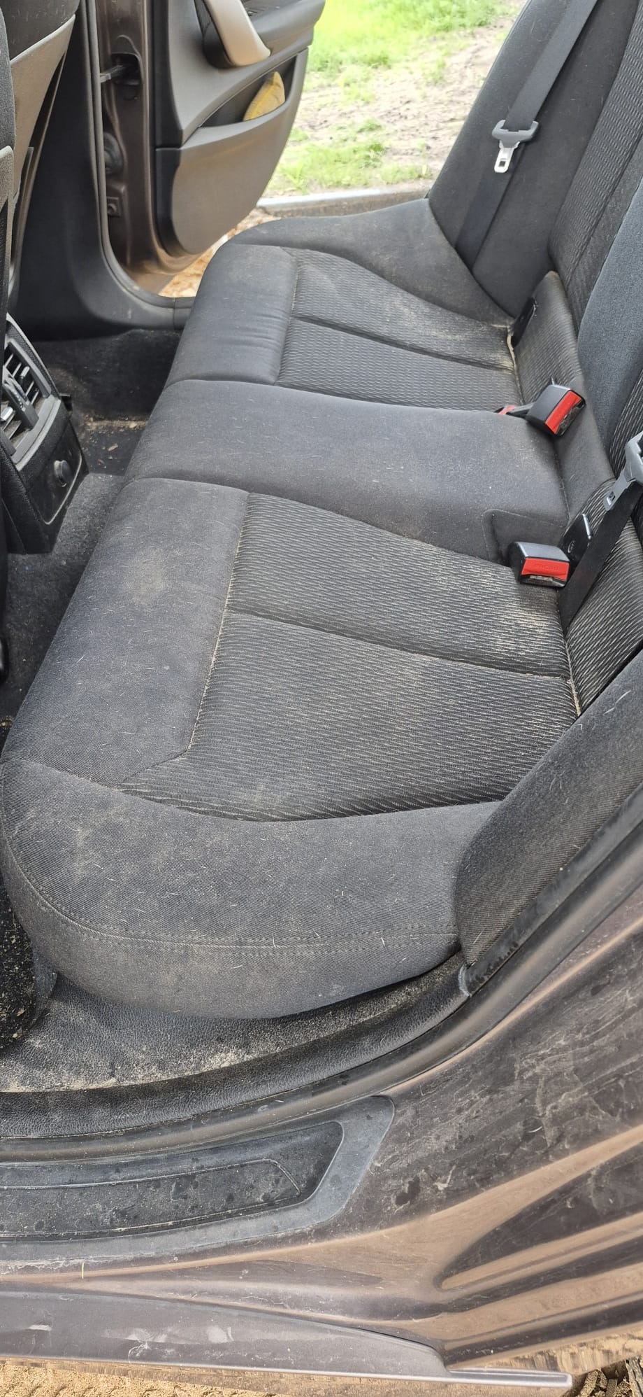 Brudna, tylna kanapa samochodowa z widocznymi plamami i zabrudzeniami na ciemnej tapicerce. Widoczne pasy bezpieczeństwa i elementy wnętrza pojazdu.