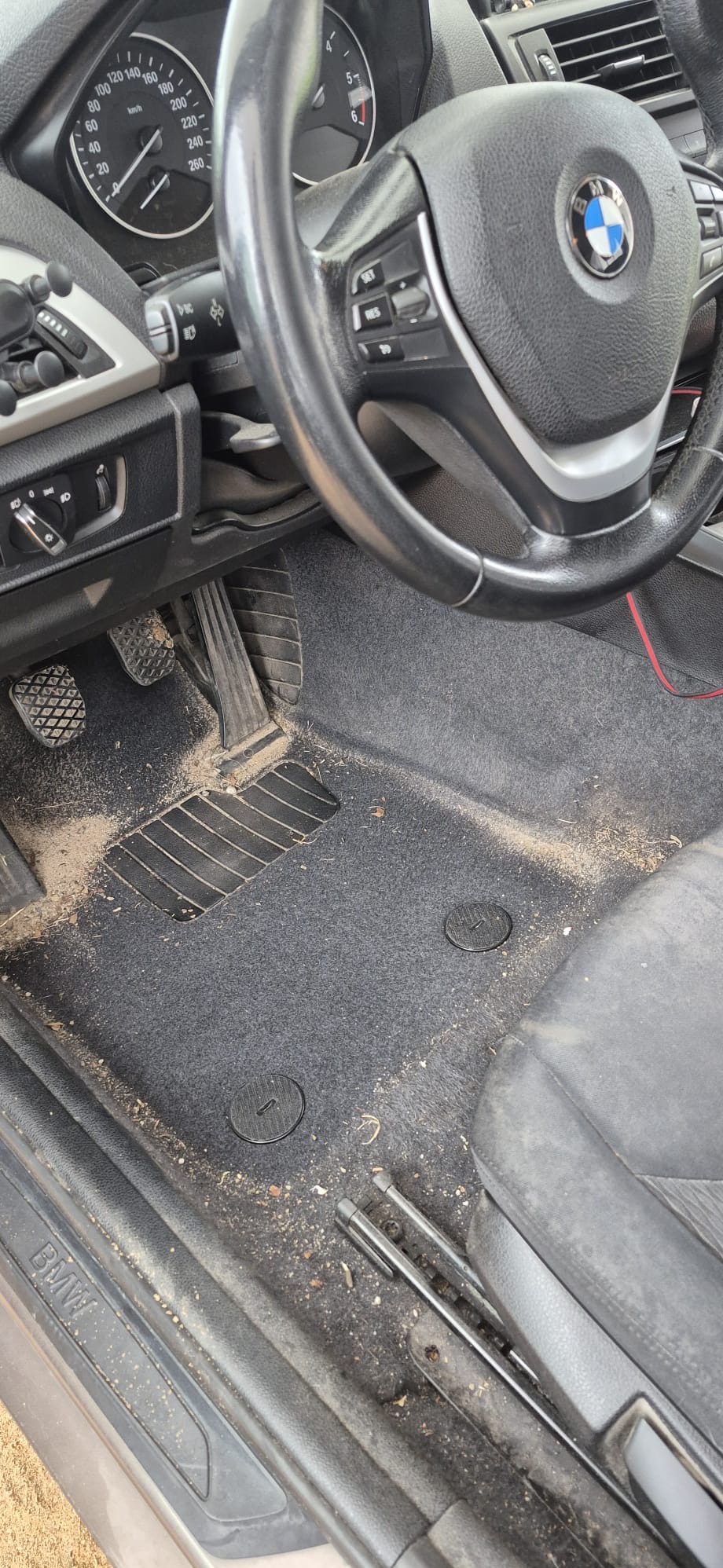 Wnętrze brudnego samochodu marki BMW. Widoczne pedały, kierownica i dywaniki pokryte piaskiem i kurzem. Potrzeba gruntownego czyszczenia tapicerki samochodowej.