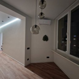 FajnaBudowa - Jasne wnętrze po wykończeniu: parkiet, białe ściany z listwami, okno z widokiem na miasto nocą, lampy wiszące i klimatyzacja. Minimalistyczny design.
