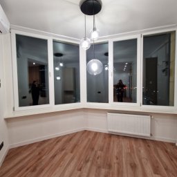 FajnaBudowa - Jasne wnętrze po remoncie: drewnopodobna podłoga, białe ściany, okno panoramiczne z widokiem na miasto, nowoczesna lampa wisząca i klimatyzator.