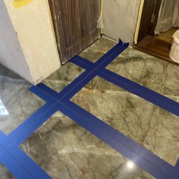 FajnaBudowa - Przygotowanie marmurowej podłogi do fugowania z użyciem niebieskiej taśmy maskującej, widoczne drzwi i fragment ściany w trakcie prac wykończeniowych.