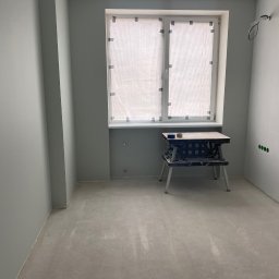 FajnaBudowa - Pokój po malowaniu na szaro, z zabezpieczonym oknem i stołem roboczym. Widoczne gniazdka elektryczne i surowa podłoga, gotowa do dalszych prac wykończeniowych.