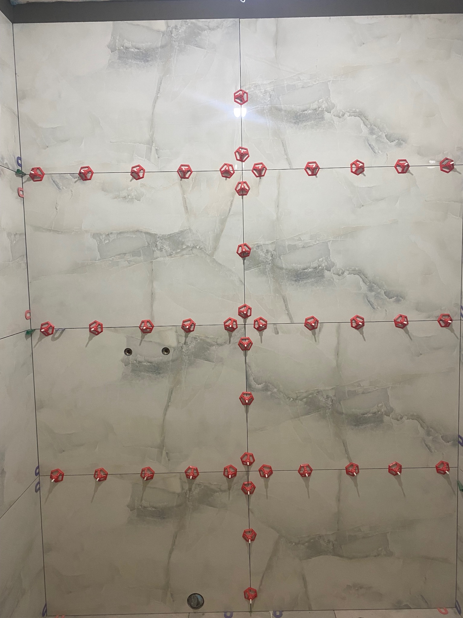 Ściana w trakcie układania płytek imitujących marmur, z użyciem czerwonych krzyżyków dystansowych i klipsów poziomujących, widoczne otwory na instalacje.