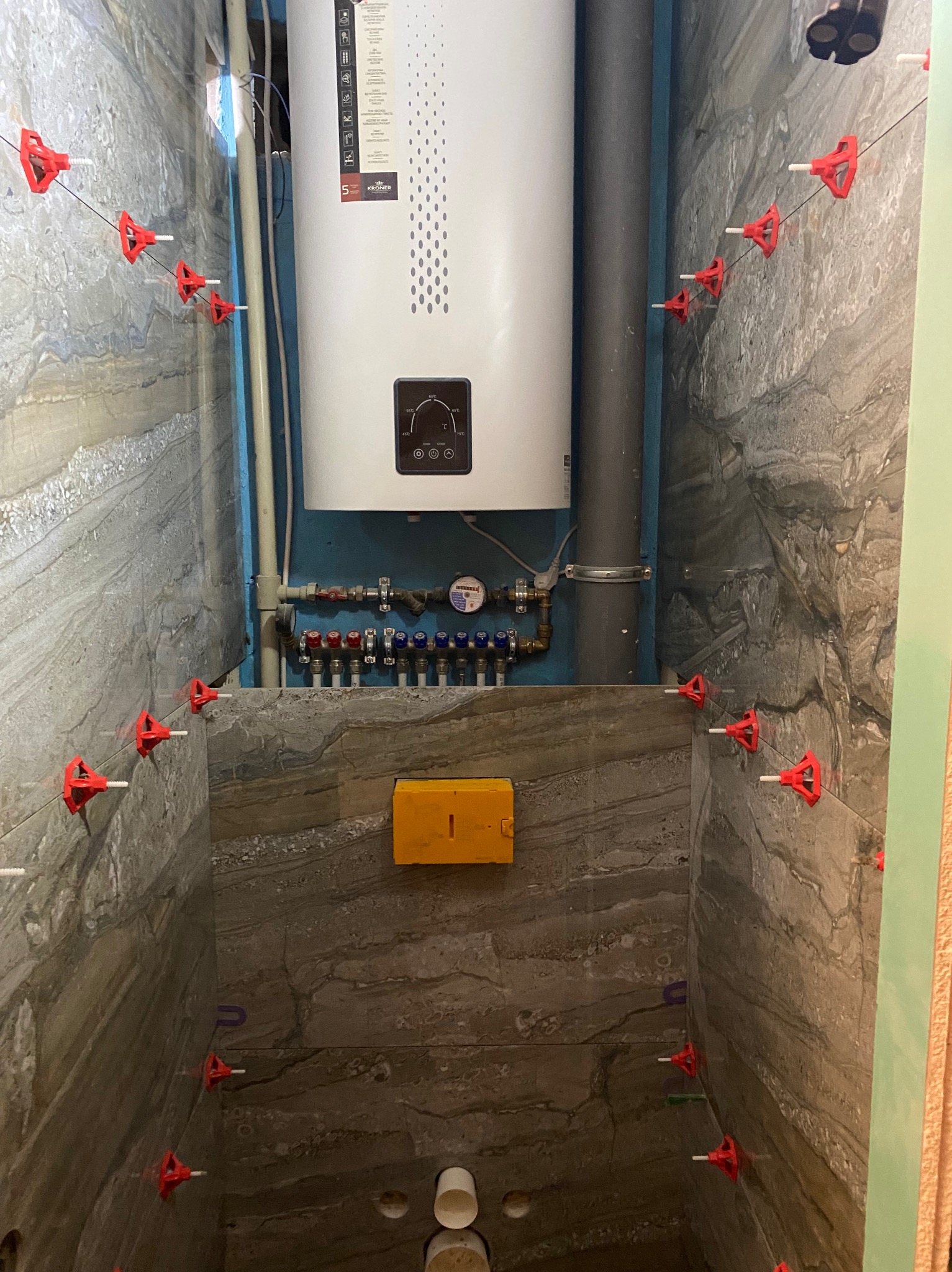 Łazienka w trakcie wykańczania: szare płytki imitujące kamień, czerwone klipsy poziomujące, bojler i instalacja wodna. Widok na fragment ściany i podłogi.