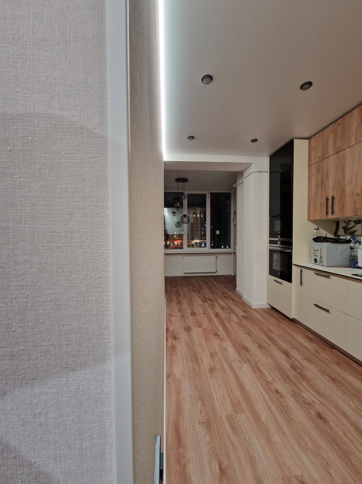 Nowoczesne wnętrze po remoncie: parkiet w odcieniach drewna, jasna kuchnia z czarnymi akcentami i widok na balkon z oświetleniem. Minimalistyczny design.