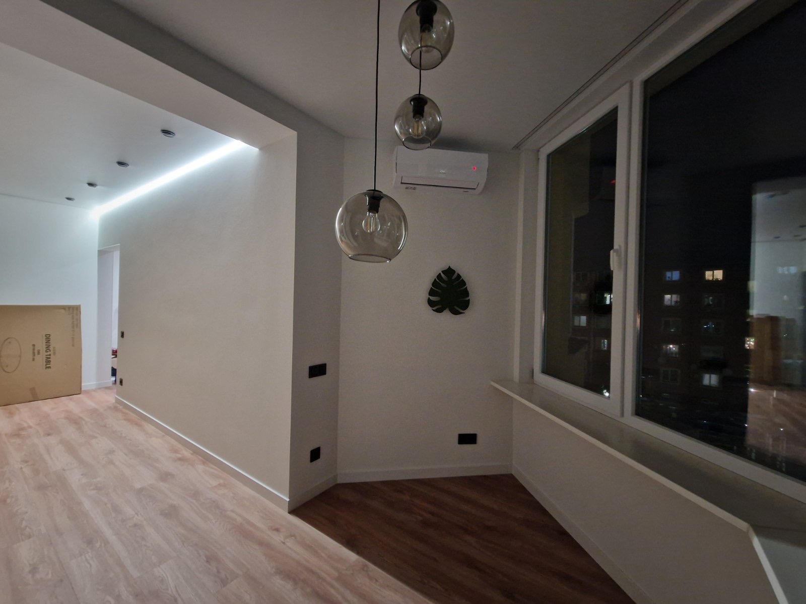 Jasne wnętrze po wykończeniu: parkiet, białe ściany z listwami, okno z widokiem na miasto nocą, lampy wiszące i klimatyzacja. Minimalistyczny design.