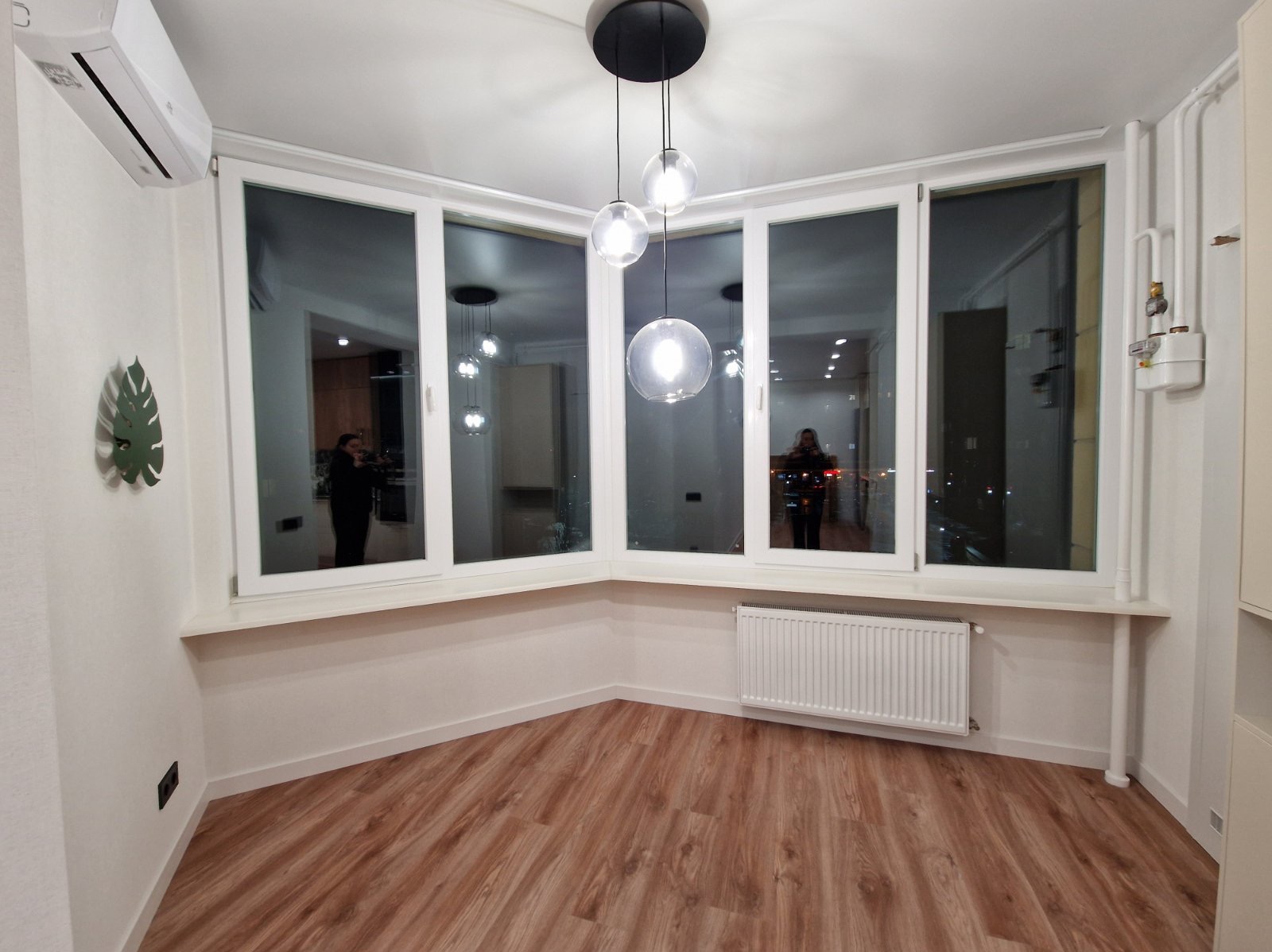 Jasne wnętrze po remoncie: drewnopodobna podłoga, białe ściany, okno panoramiczne z widokiem na miasto, nowoczesna lampa wisząca i klimatyzator.