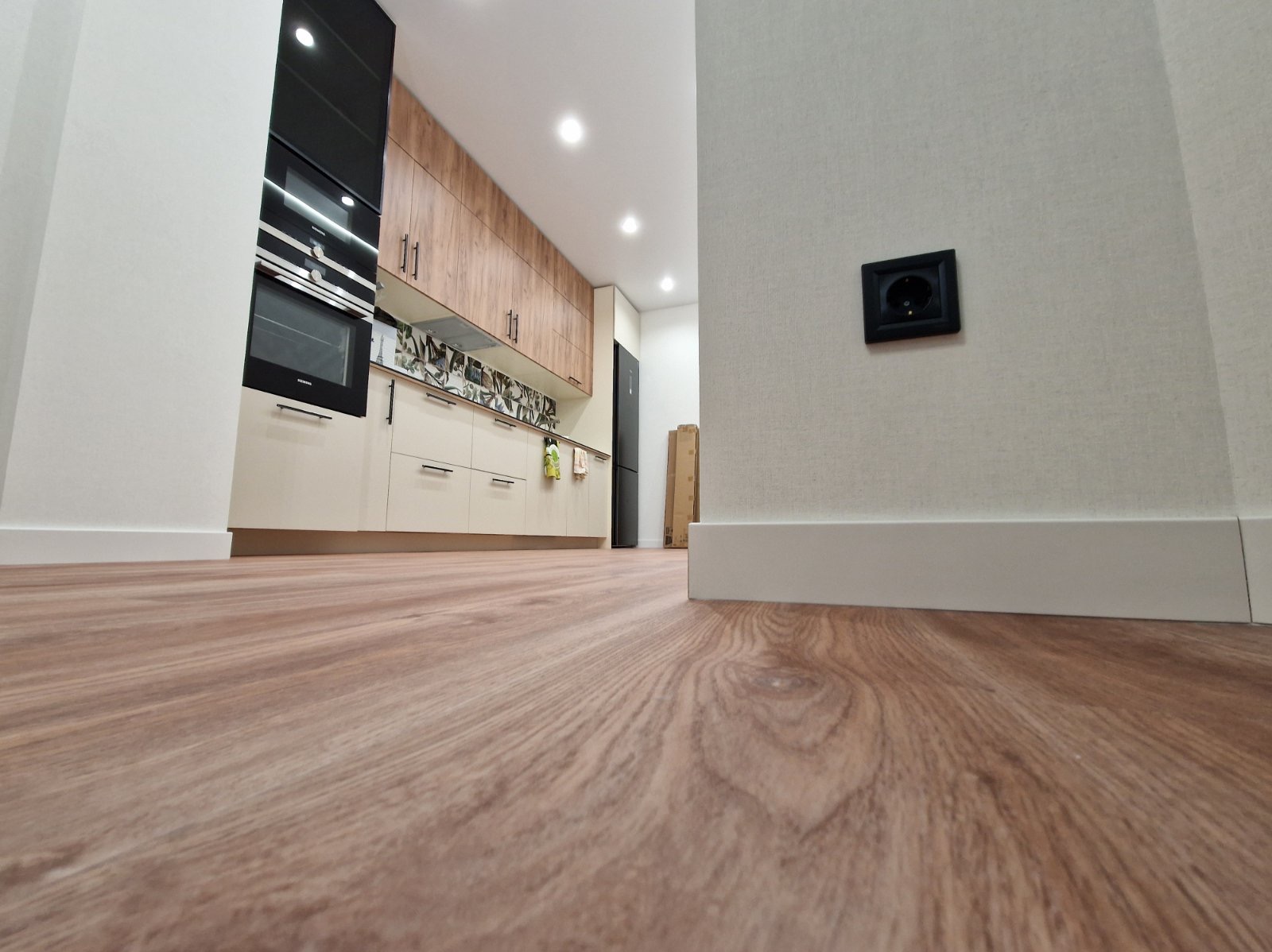 Nowoczesna kuchnia z drewnianą podłogą i białymi listwami przypodłogowymi. Na ścianie czarne gniazdko elektryczne. Ujęcie z niskiej perspektywy.