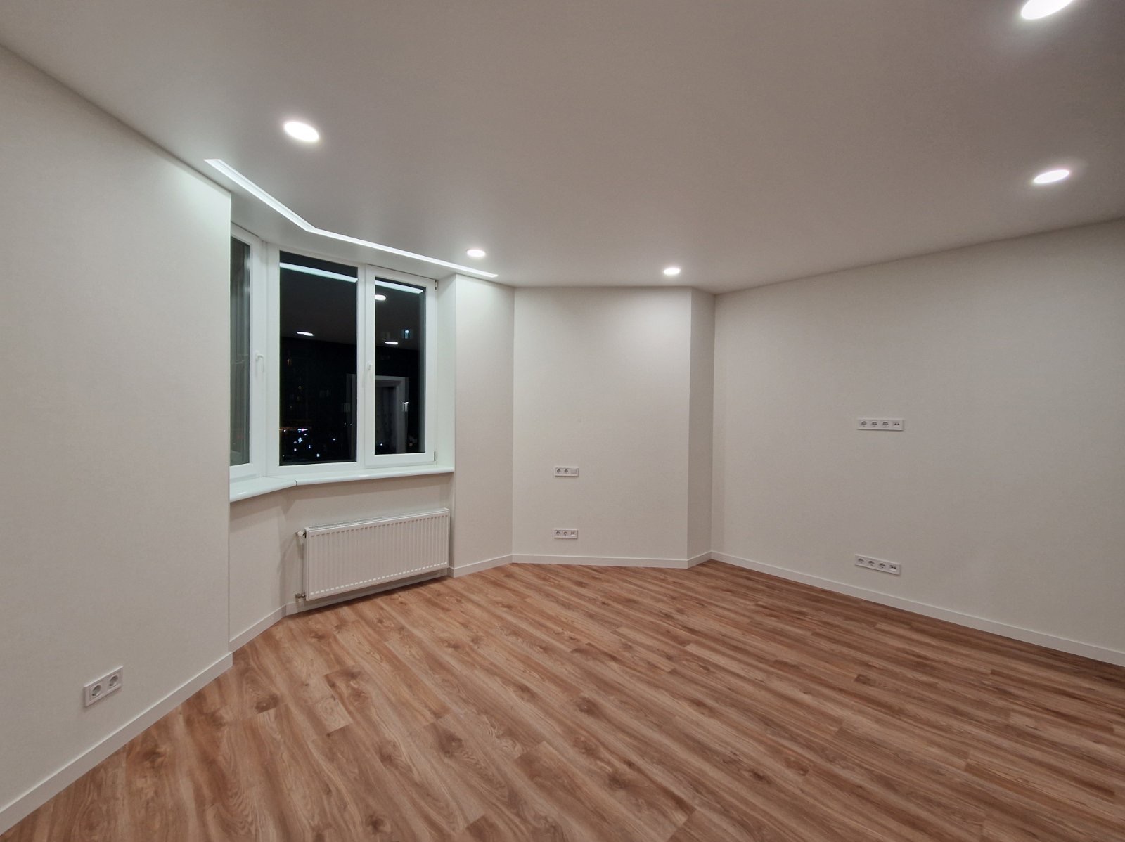 Jasne, odnowione wnętrze pokoju z drewnianą podłogą, białymi ścianami i oknem wykuszowym. Widoczne gniazdka elektryczne i grzejnik. Nowoczesne oświetlenie sufitowe.