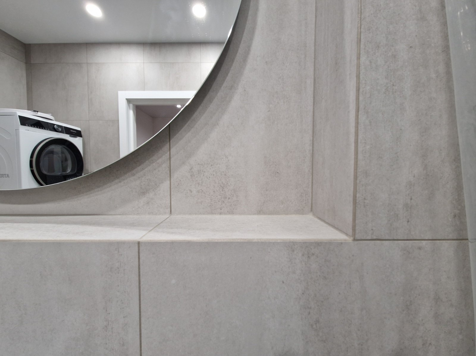 Szare, matowe płytki w łazience z okrągłym lustrem, w którym odbija się pralka i drzwi. Minimalistyczny design, neutralna kolorystyka, nowoczesne wykończenie.