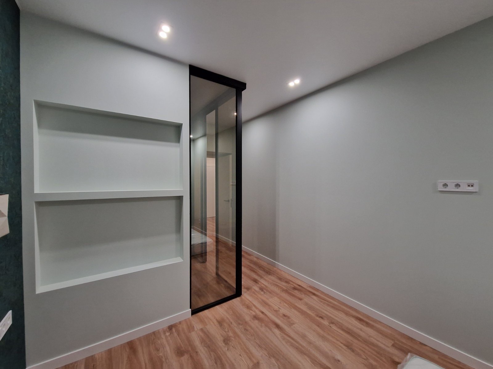 Jasne wnętrze po wykończeniu: drewnopodobna podłoga, ściany w odcieniach szarości z wnękami i gładkimi powierzchniami, szklane drzwi z czarną ramą.