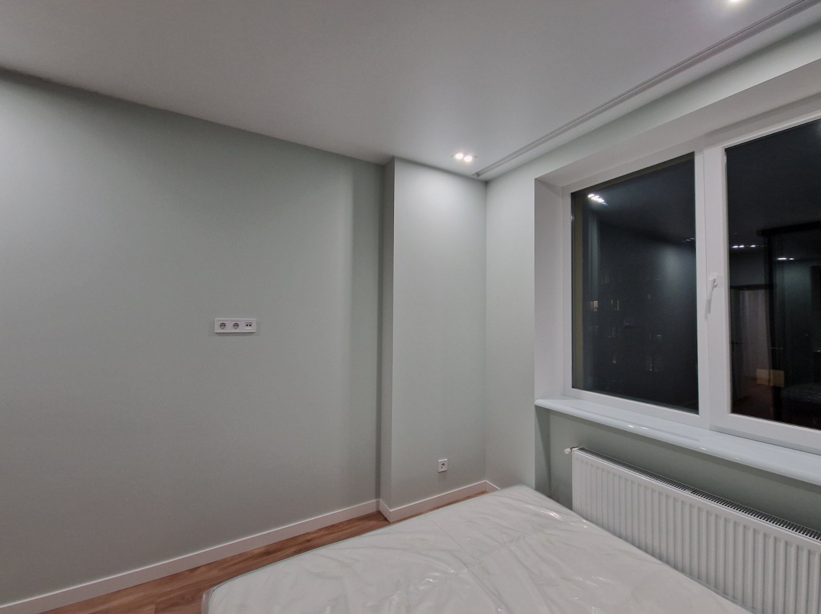 Jasne wnętrze pokoju po malowaniu ścian na odcień szarości, z widocznym grzejnikiem pod oknem i częściowo zakrytym łóżkiem folią ochronną.