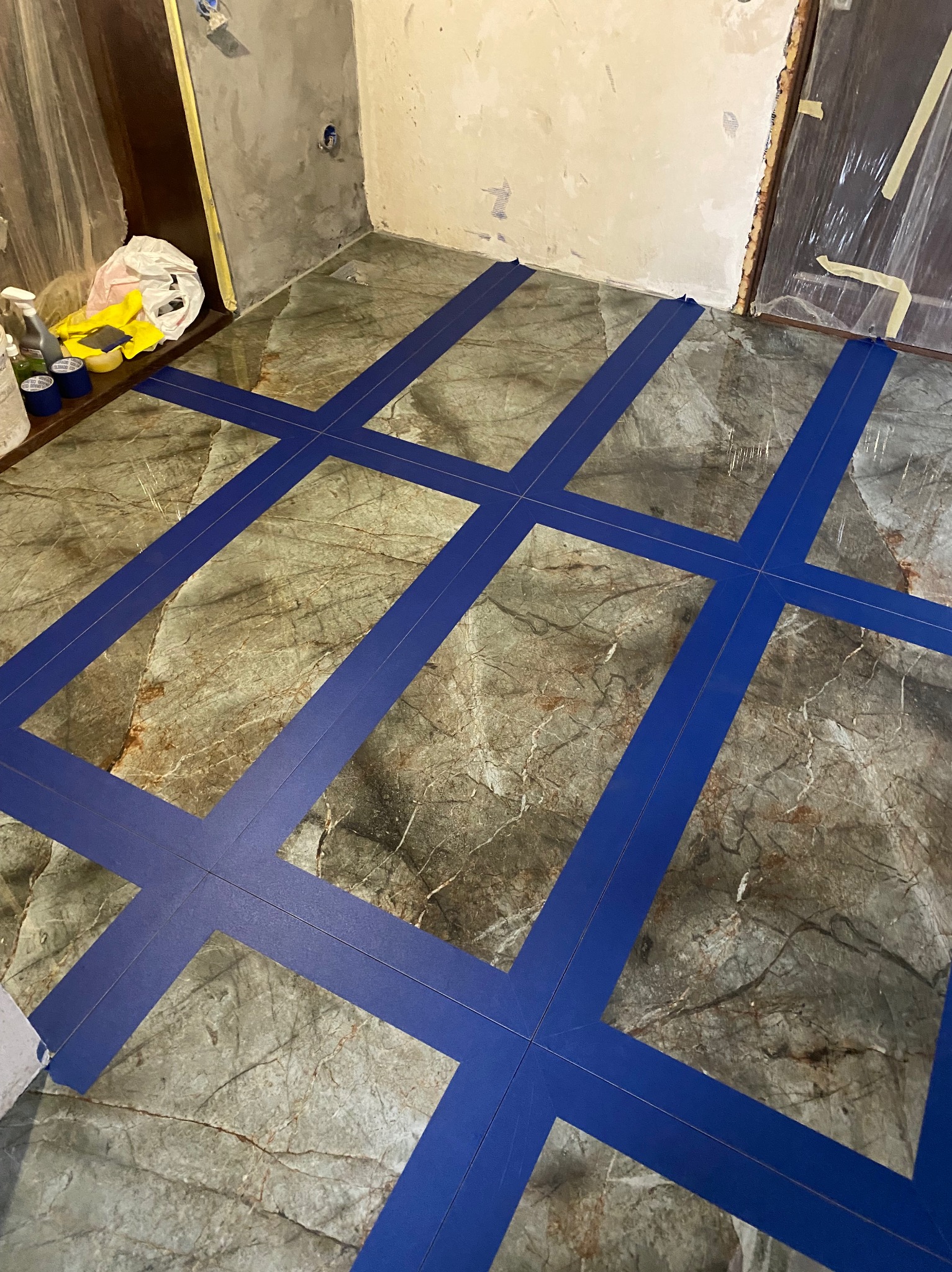 Przygotowanie do malowania wzoru na marmurowej podłodze w łazience, wyklejonej niebieską taśmą malarską. Widoczne narzędzia i materiały malarskie.