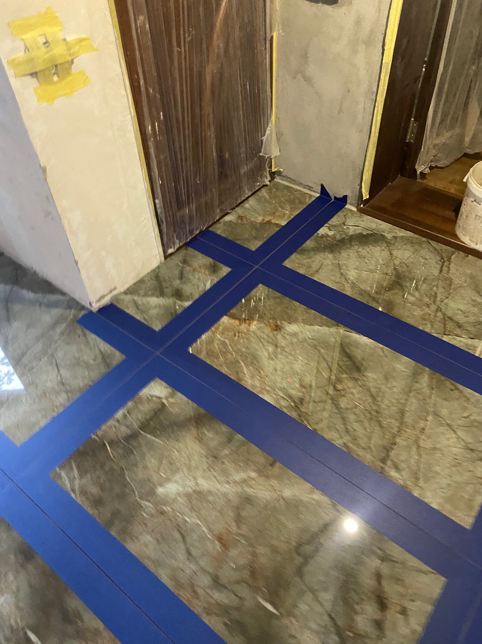 Przygotowanie marmurowej podłogi do fugowania z użyciem niebieskiej taśmy maskującej, widoczne drzwi i fragment ściany w trakcie prac wykończeniowych.