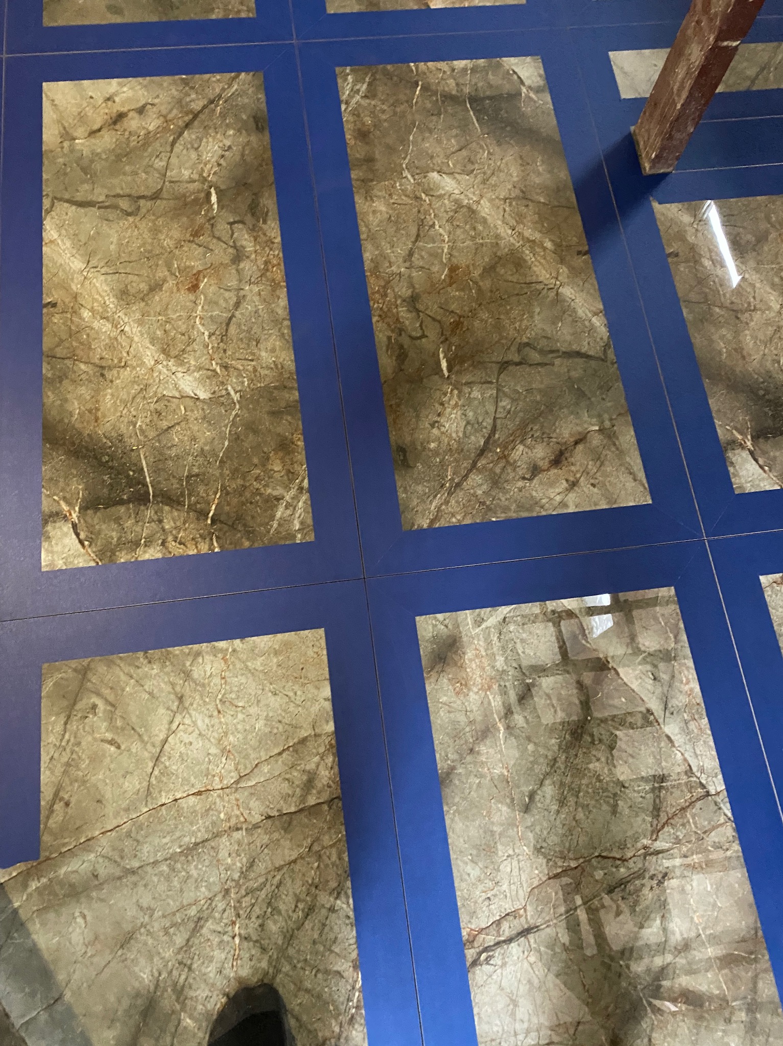 Ułożona podłoga z płytek imitujących marmur, z wyraźnymi, niebieskimi fugami. Fragment drabiny w prawym górnym rogu. Widoczne odbicie światła na powierzchni.