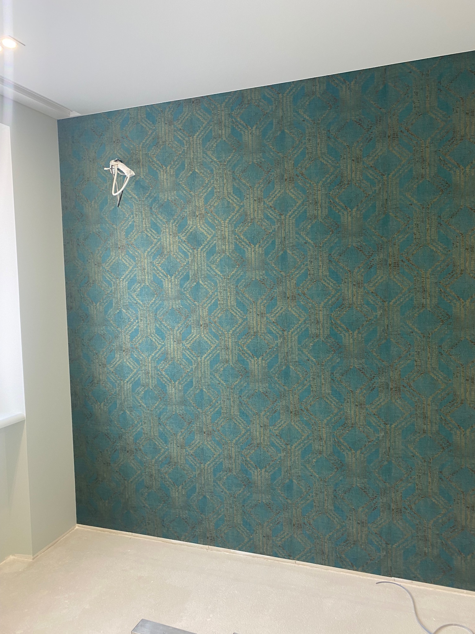 Ściana z tapetą w geometryczny wzór w odcieniach turkusu i złota. Widoczne kable i fragment sufitu z oświetleniem. Świeżo wykończone wnętrze.