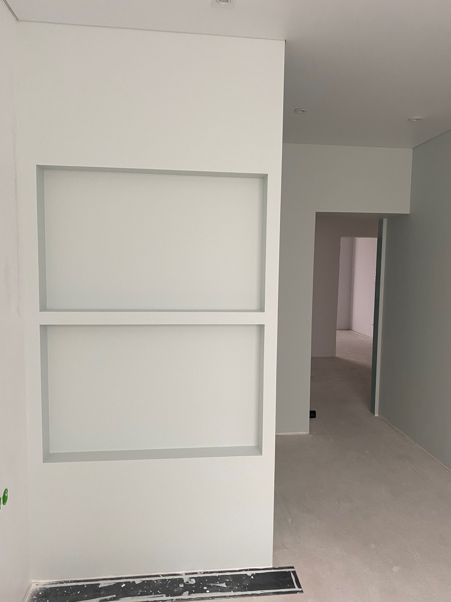 Minimalistyczne wnętrze po malowaniu: białe ściany z wnękami i szara podłoga. Widok na korytarz z drzwiami. Oświetlenie sufitowe.