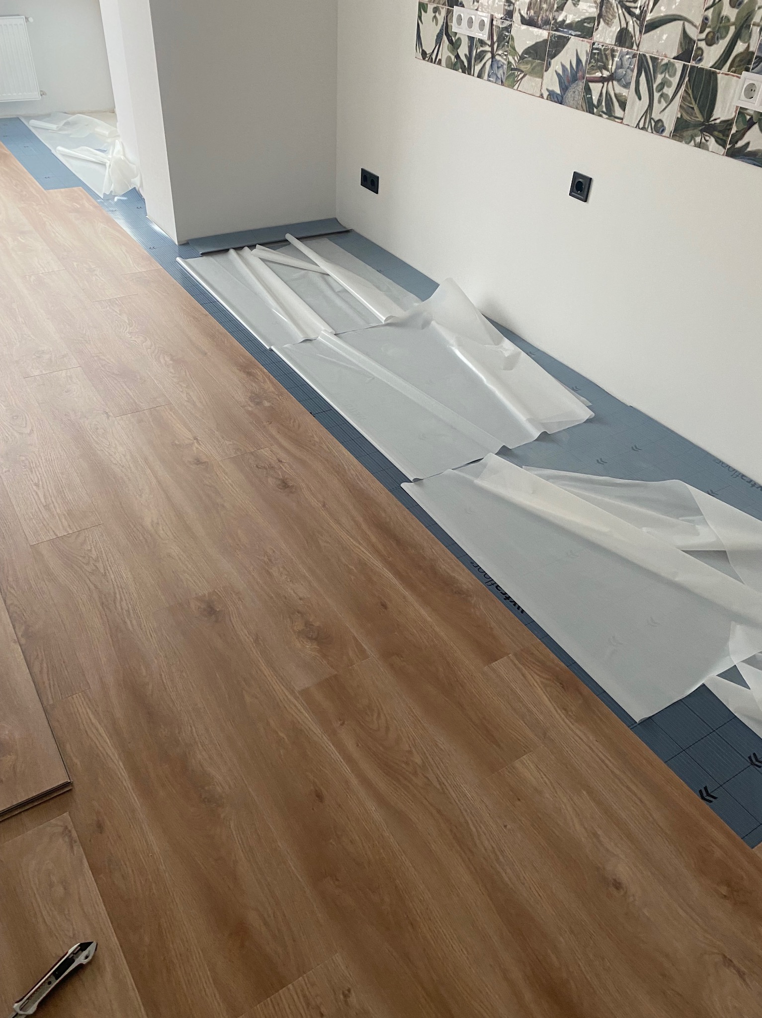 Układanie paneli podłogowych w jasnym wnętrzu. Częściowo rozłożona folia ochronna na podkładzie. W tle biała ściana z dekoracyjnymi płytkami i gniazdkami.