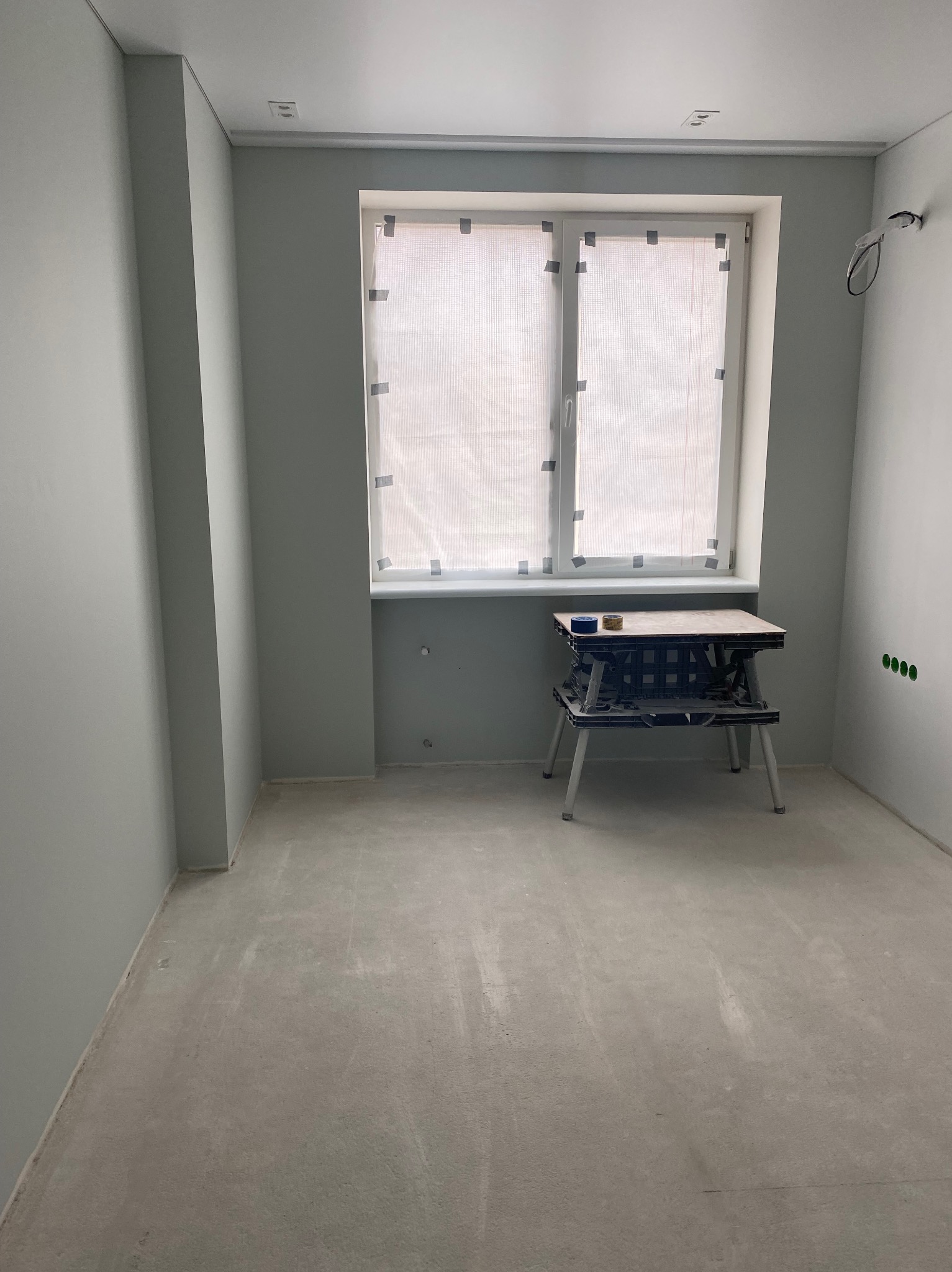 Pokój po malowaniu na szaro, z zabezpieczonym oknem i stołem roboczym. Widoczne gniazdka elektryczne i surowa podłoga, gotowa do dalszych prac wykończeniowych.