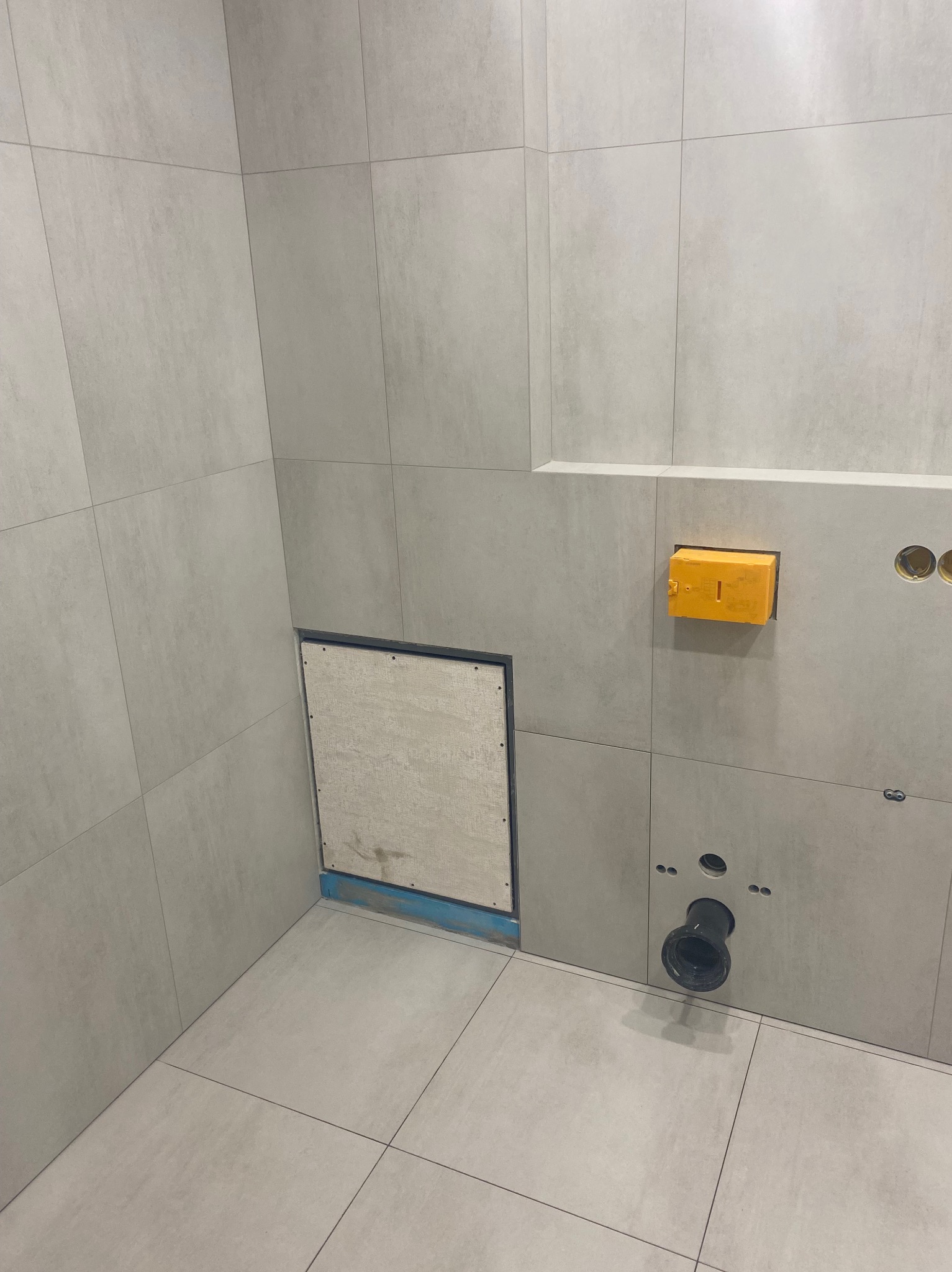 Wykończona łazienka z szarymi płytkami, widoczne otwory na instalacje i rewizja. Minimalistyczny design, surowy styl.