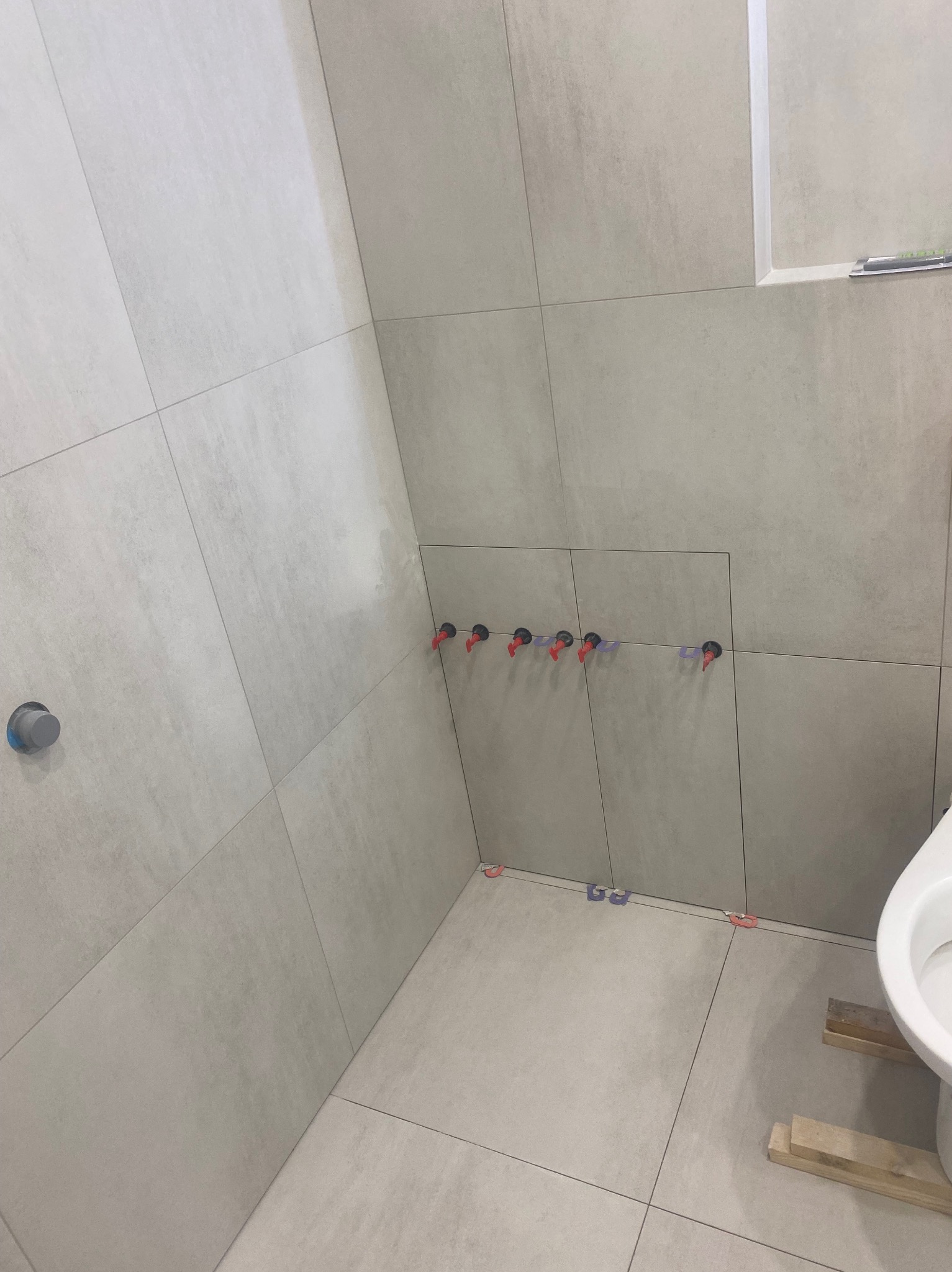 Jasne, kwadratowe płytki na ścianach i podłodze łazienki w trakcie montażu. Widoczne krzyżyki dystansowe i otwory na rury z czerwonymi elementami mocującymi.