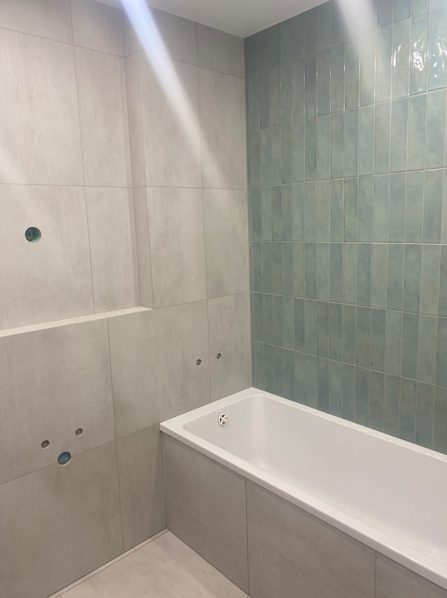 Wykończona łazienka w Bydgoszczy z wanną i częściowo ułożonymi płytkami w odcieniach szarości i błękitu. Widoczne otwory na armaturę.