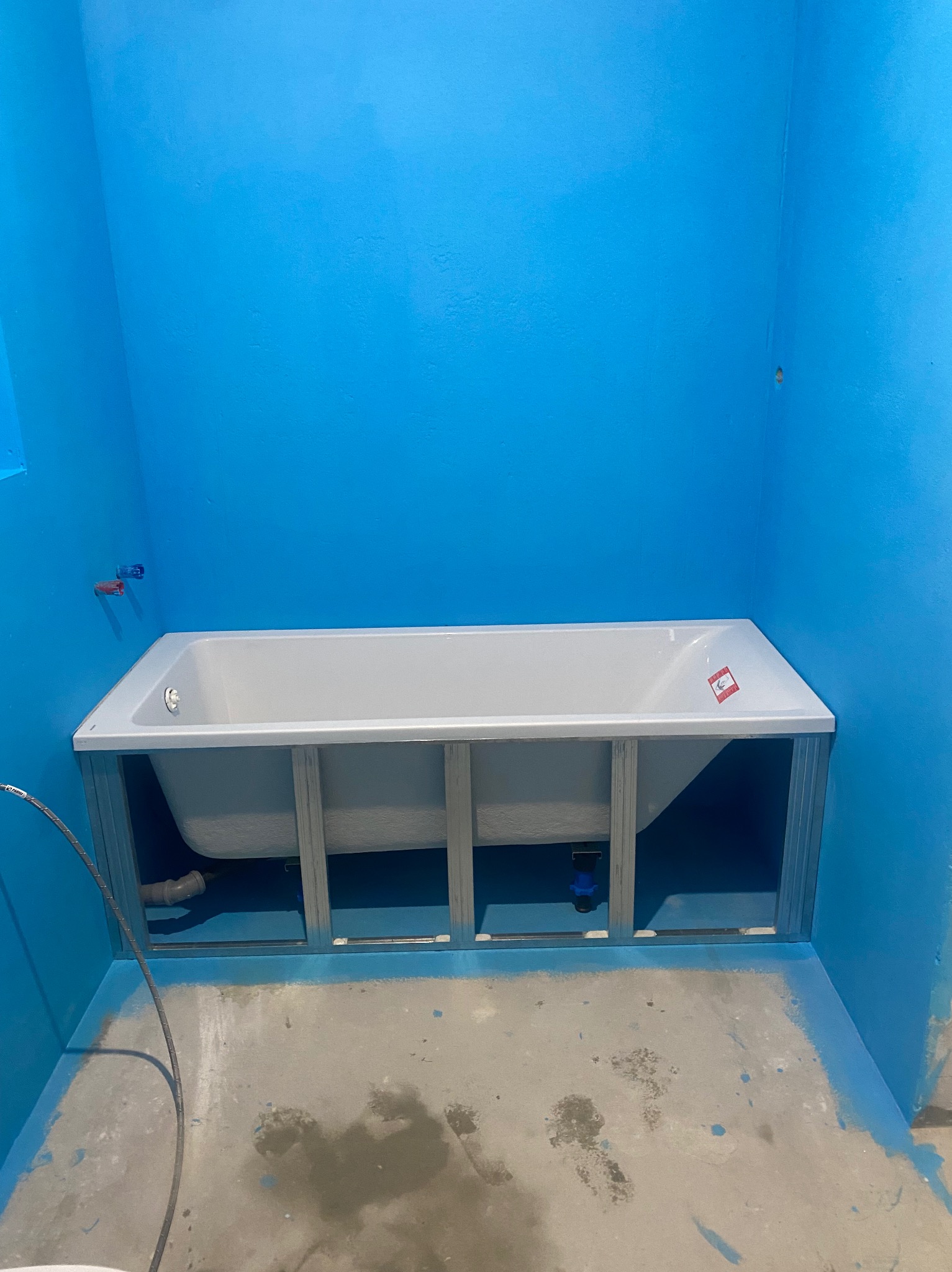 Biała wanna obudowana metalową konstrukcją w łazience z niebieskimi ścianami i podłogą pokrytą hydroizolacją. Widoczne elementy instalacji wodnej.