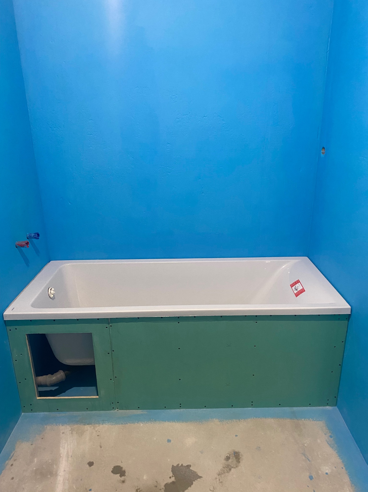 Niebieska łazienka w trakcie wykończenia: biała wanna obudowana zielonymi płytami gipsowymi, widoczne rury. Ściany pomalowane na intensywny, błękitny kolor.