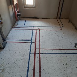 Instal Plus Michał Potrzeba - Instalacja wodna TECEflex z czerwono-niebieskimi rurami ułożonymi na styropianie izolacyjnym w surowym wnętrzu łazienki z oknem i stelażem podtynkowym WC.