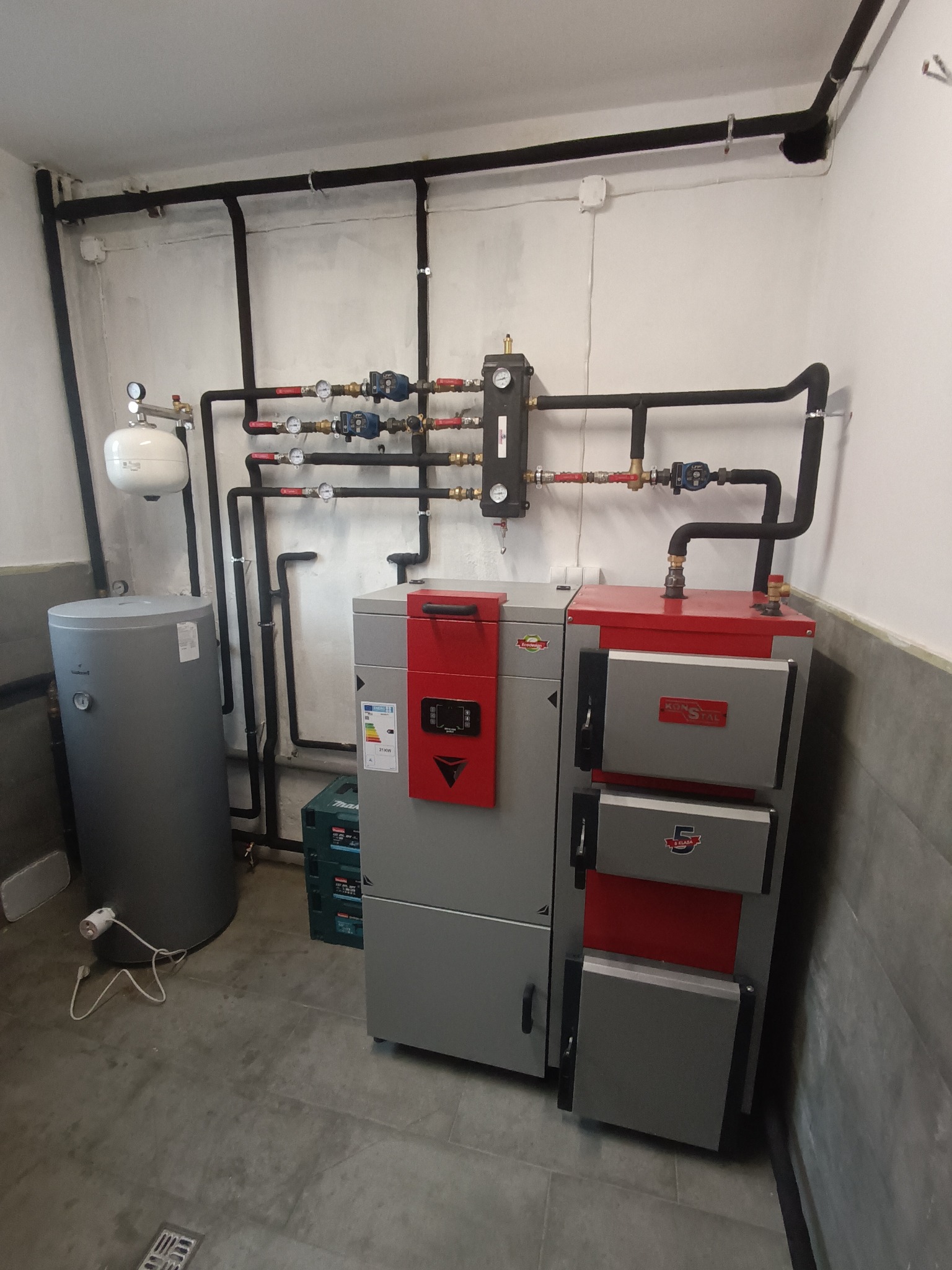 Kompaktowa kotłownia pelletowa KonStal z szarym zbiornikiem i czarnymi rurami, zainstalowana w pomieszczeniu z szarą podłogą i białymi ścianami.