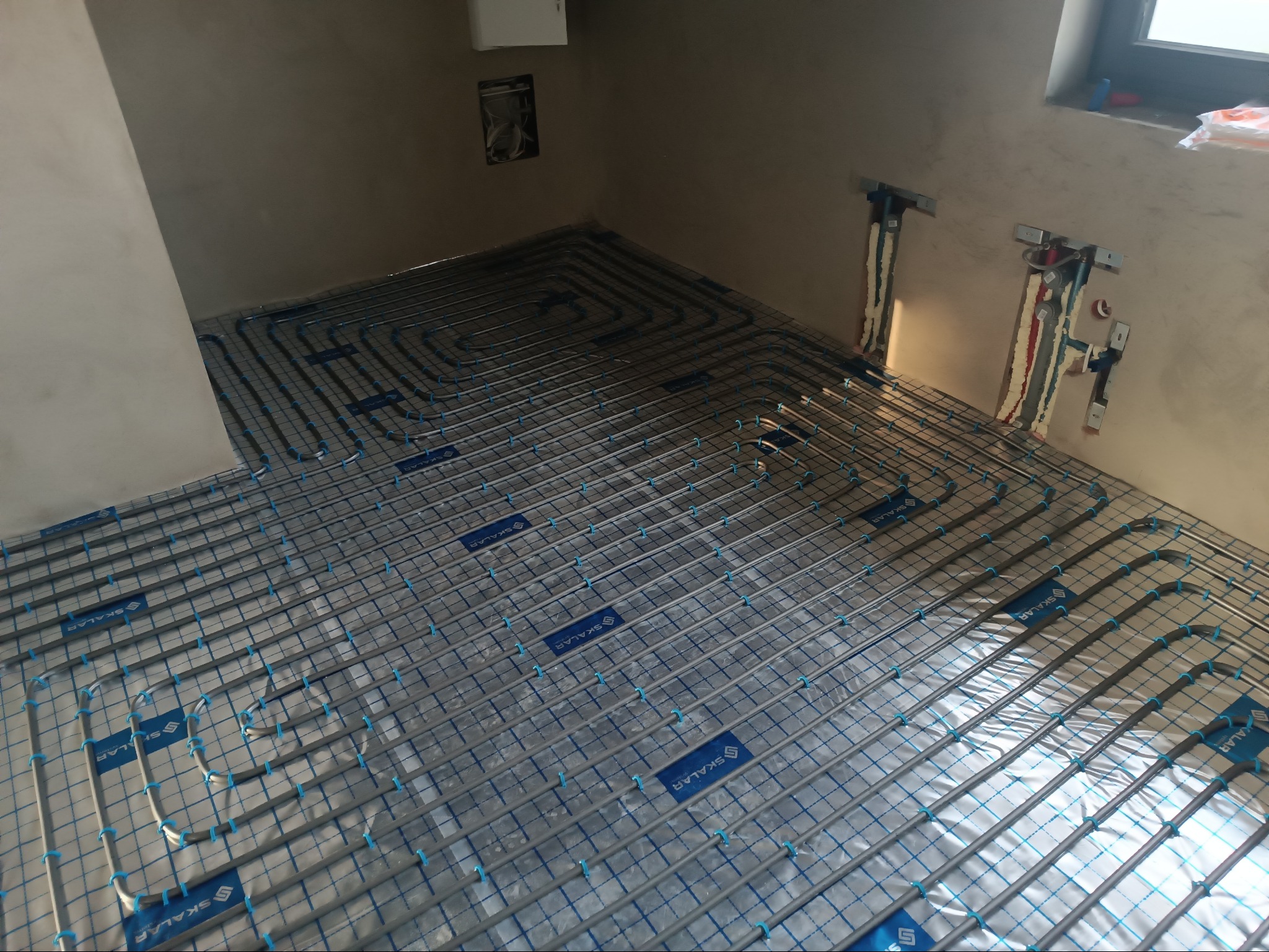 Szare rury ogrzewania podłogowego ułożone na folii z niebieską siatką w surowym wnętrzu. Widoczne przyłącza na ścianie. System Tece flex.