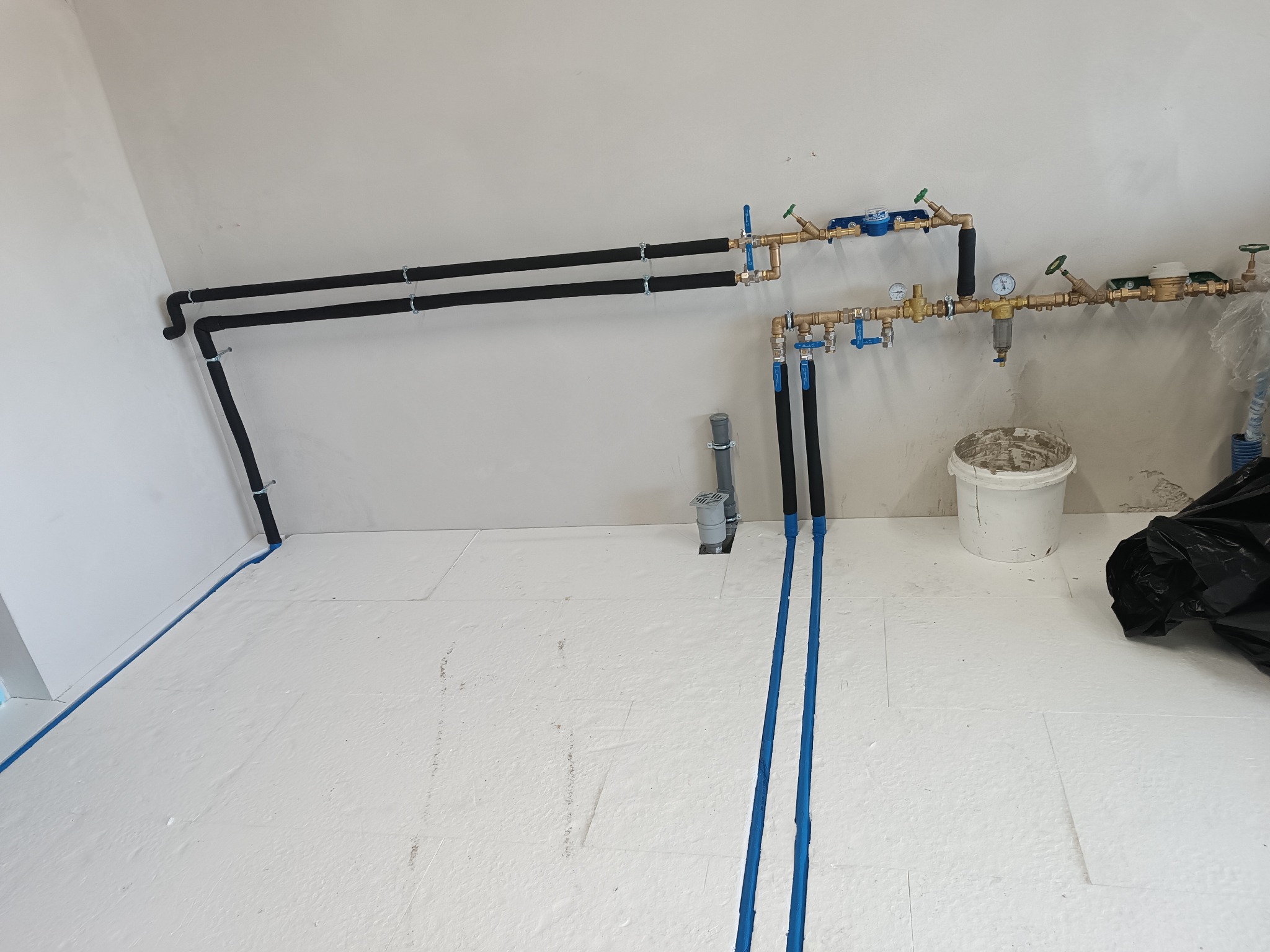 Biała podłoga z izolacją, nieotynkowane ściany i rury CO/wod-kan z zaworami, manometrami i filtrem, zamontowane przy ścianie. Widoczny fragment instalacji.