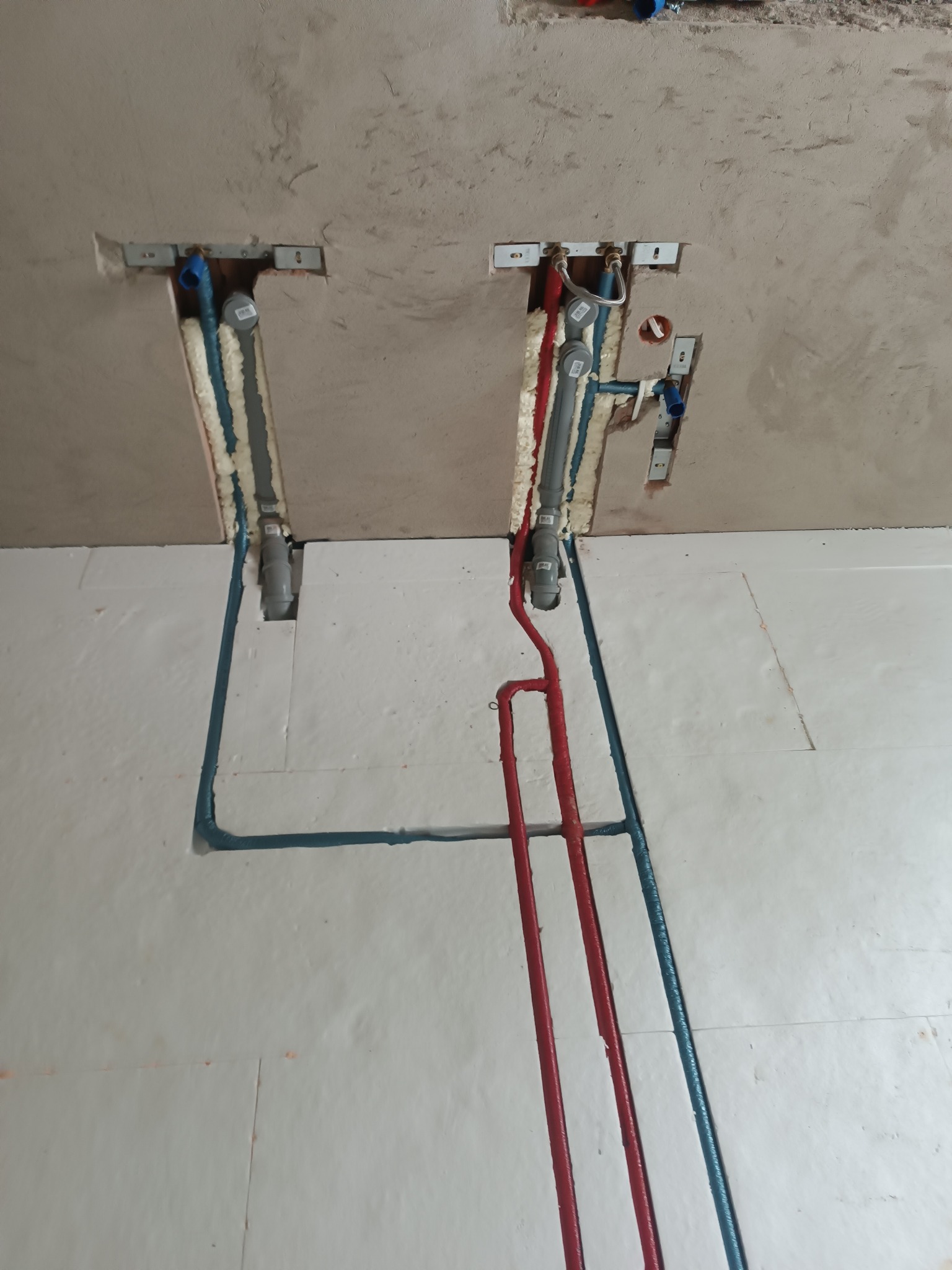 Instalacja wodna TECEflex: czerwone i niebieskie rury w izolacji styropianowej, prowadzone w ścianie i podłodze. Widoczne mocowania i pianka montażowa.