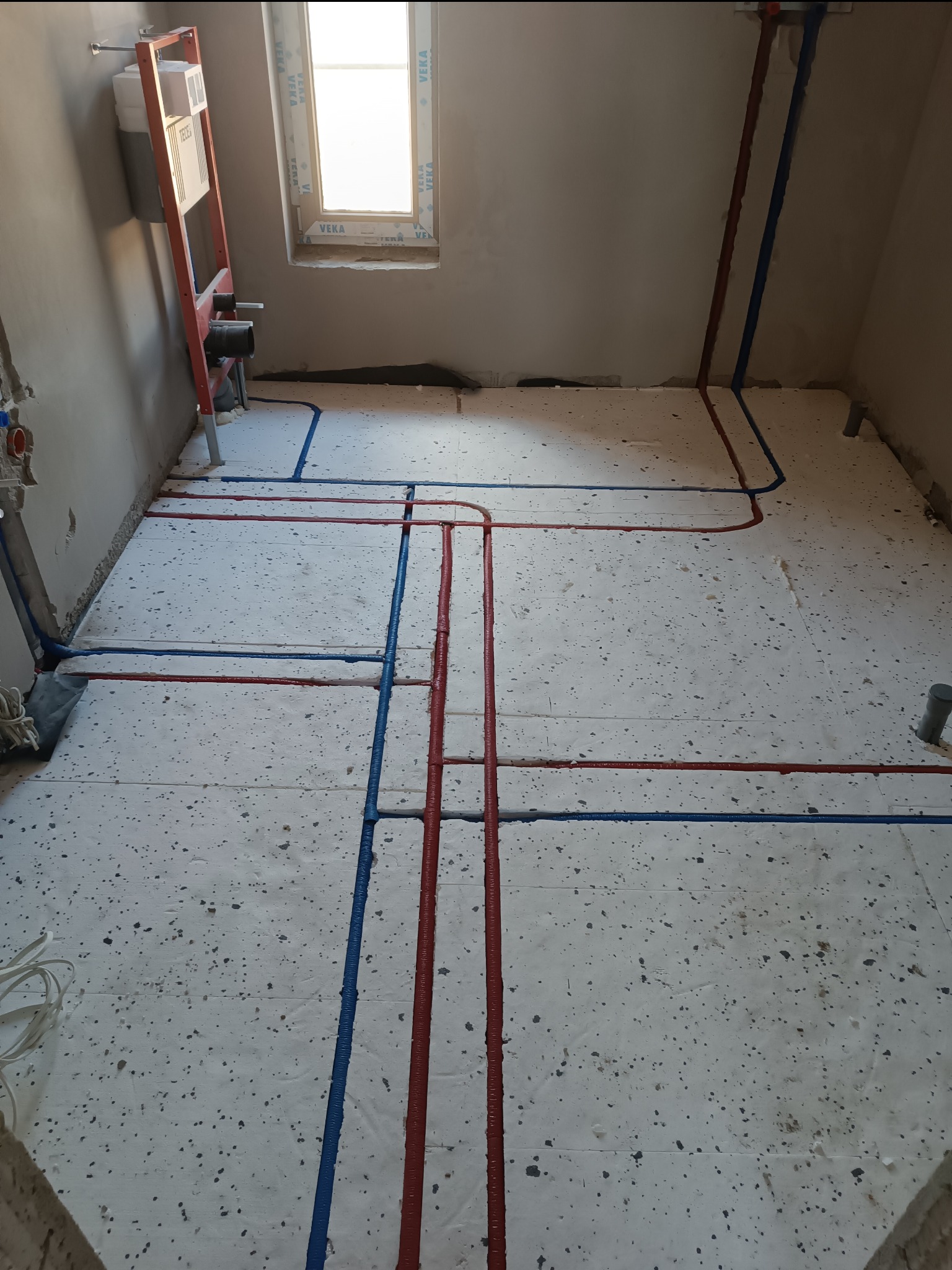 Instalacja wodna TECEflex z czerwono-niebieskimi rurami ułożonymi na styropianie izolacyjnym w surowym wnętrzu łazienki z oknem i stelażem podtynkowym WC.