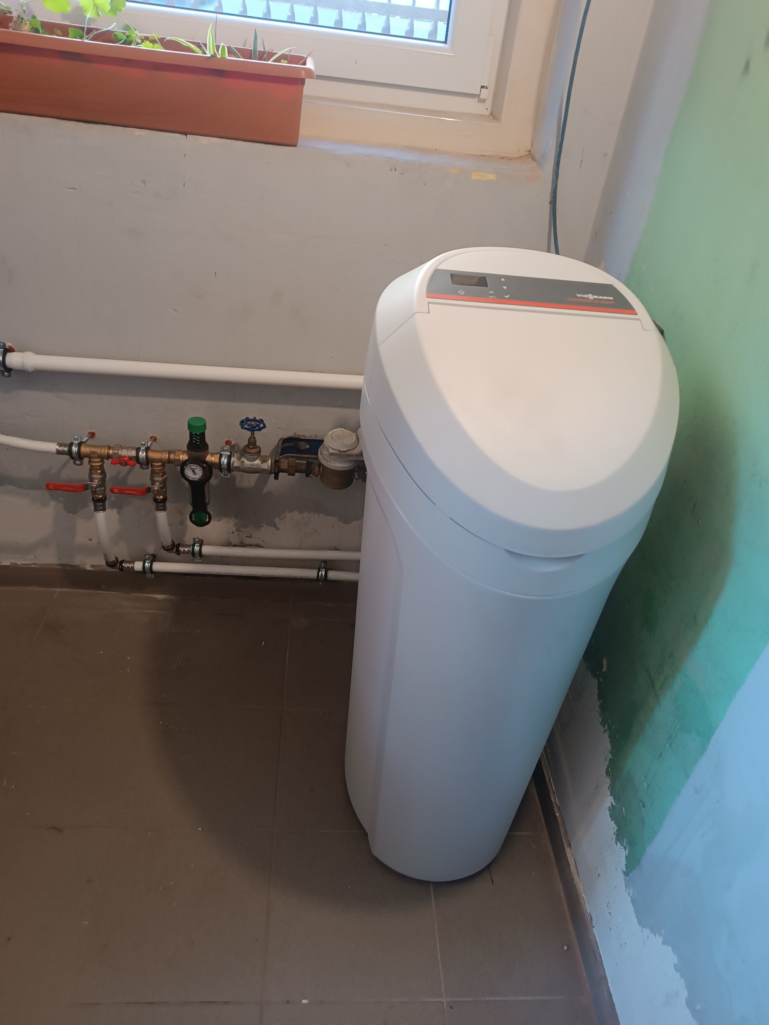 Biała stacja uzdatniania wody Viessmann Aqua Home 20, z podłączonymi rurami, stoi w rogu pomieszczenia z oknem i kwiatami. Widoczne przyłącza hydrauliczne.