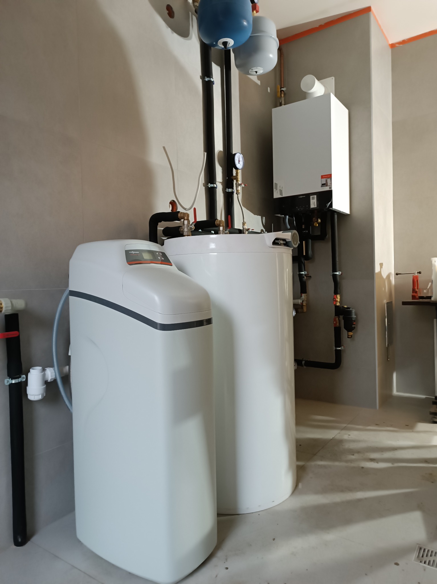 Nowoczesna kotłownia z białym piecem gazowym Viessmann, zasobnikiem wody i stacją uzdatniania. Czarno-biała instalacja, szare płytki na ścianach i podłodze.