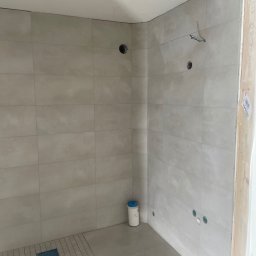 Angel-bud - Wykończona łazienka z jasnymi płytkami, widoczne otwory na armaturę i odpływ liniowy. Minimalistyczny design, surowy stan przed montażem wyposażenia.
