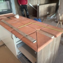 Angel-bud - Montaż terakoty na blacie kuchennym. Widoczne poziomica, miarka i białe szafki. W tle fragment pieca i osoba w czerwonej bluzie. Ujęcie z bliska, w trakcie prac.