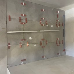 Angel-bud - Ściana w trakcie układania szarych płytek, z użyciem poziomicy i krzyżyków dystansowych. Widoczne fragmenty podłogi wyłożonej jasnymi płytkami.