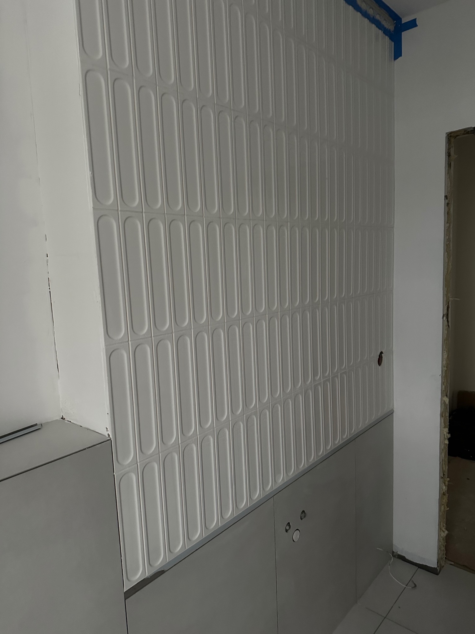 Białe, podłużne płytki ułożone pionowo na ścianie, dolna część ściany wykończona szarymi panelami. Widoczne fragmenty instalacji elektrycznej i niebieska taśma malarska.
