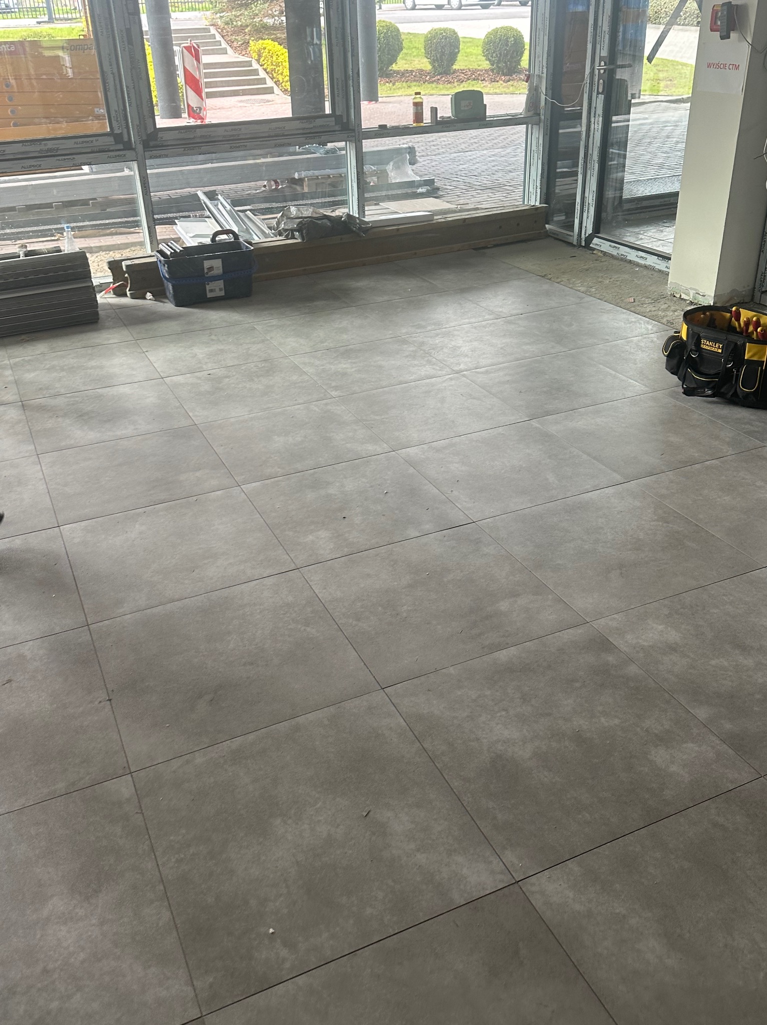 Ułożone, szare płytki podłogowe w nowym pomieszczeniu z dużymi oknami. Widoczny kufer narzędziowy i torba narzędziowa.