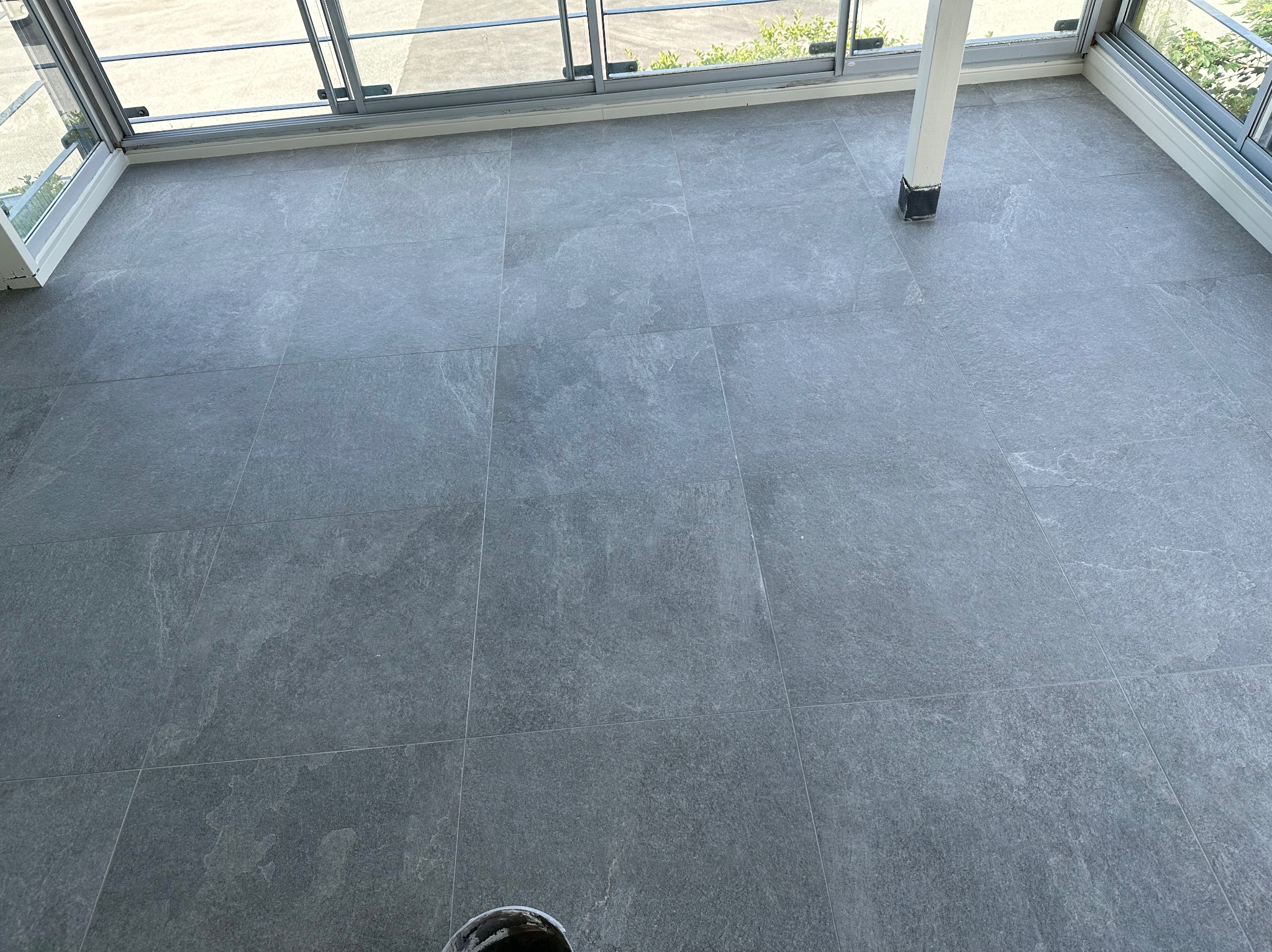 Szare, matowe płytki na podłodze balkonu z widocznymi szklanymi drzwiami balkonowymi i fragmentem kolumny. Minimalistyczny design, równomierne ułożenie płytek.