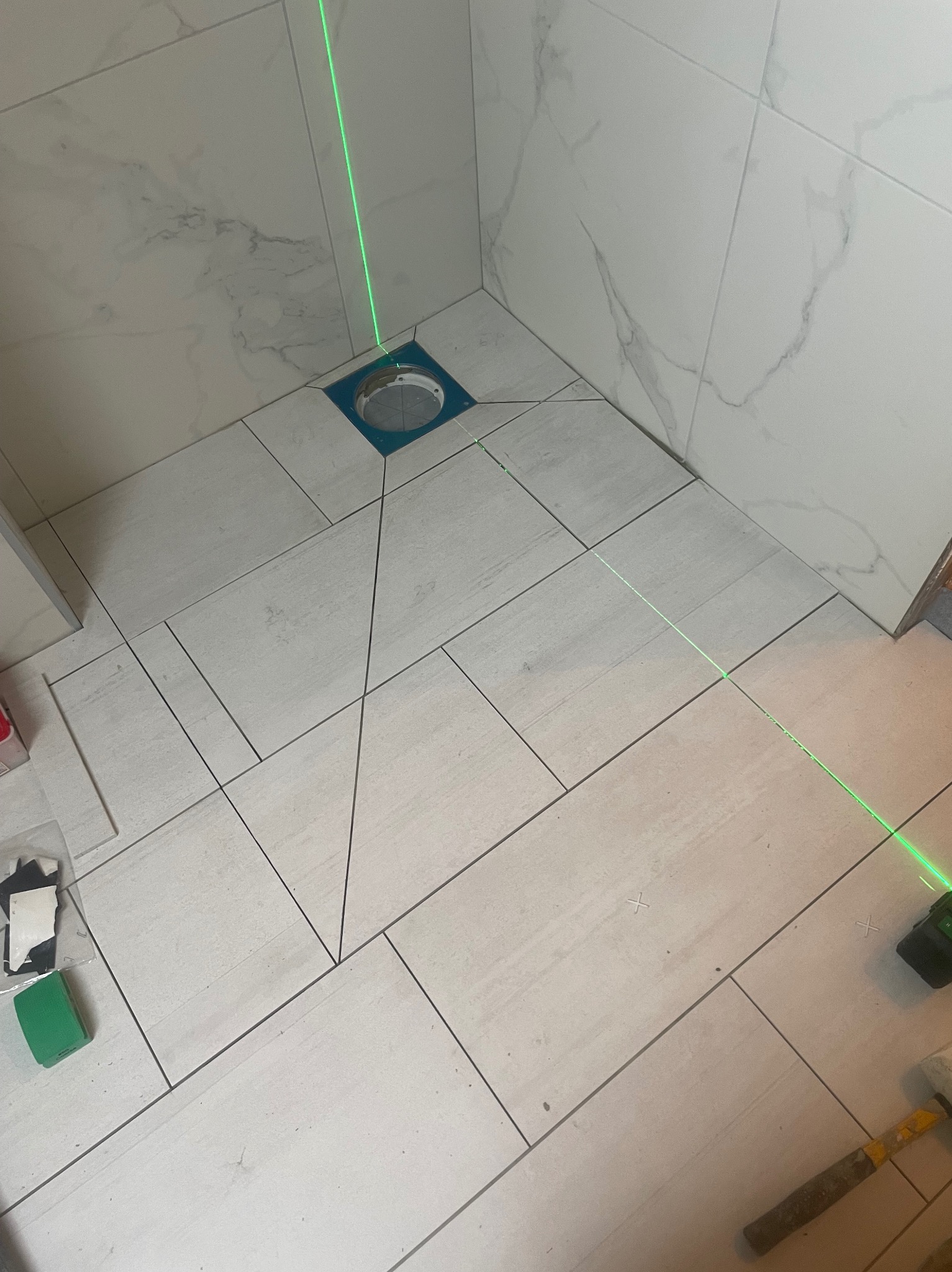 Układanie płytek podłogowych w łazience, z użyciem lasera krzyżowego do precyzyjnego wyznaczania linii. Widoczny odpływ liniowy i narzędzia glazurnicze.