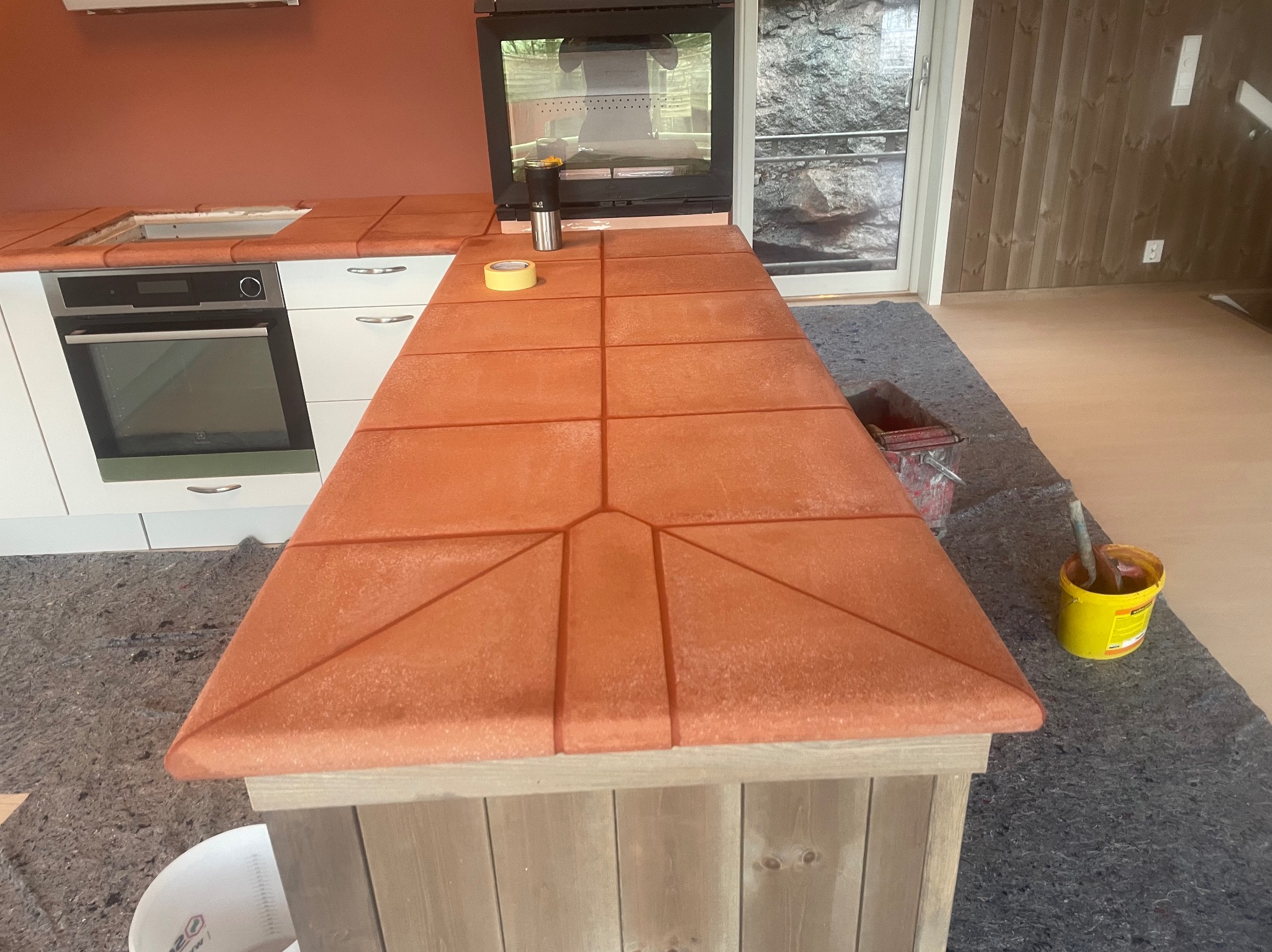 Pomarańczowa, kafelkowana wyspa kuchenna z unikalnym wzorem, widoczna na tle białych szafek i okna. Na blacie taśma malarska i akcesoria. W tle drewniana ściana.