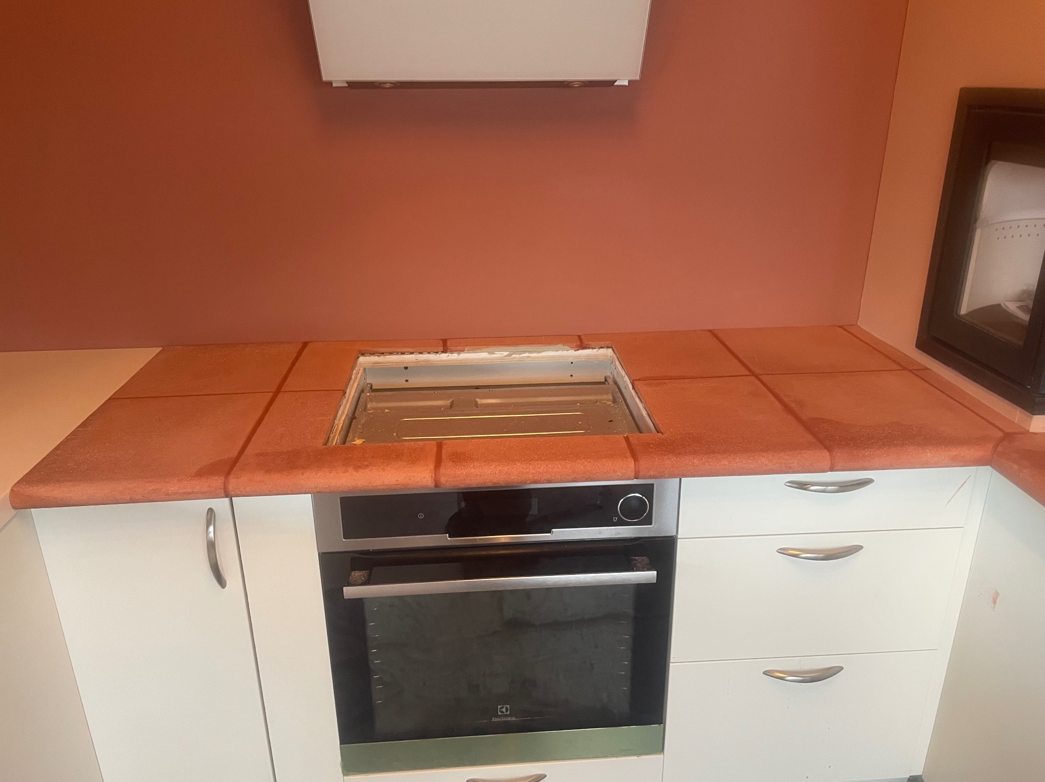 Kuchnia po remoncie: terakotowy blat z wycięciem na płytę grzewczą nad piekarnikiem Electrolux, białe szafki, ściana w odcieniach terakoty, nowoczesny okap.