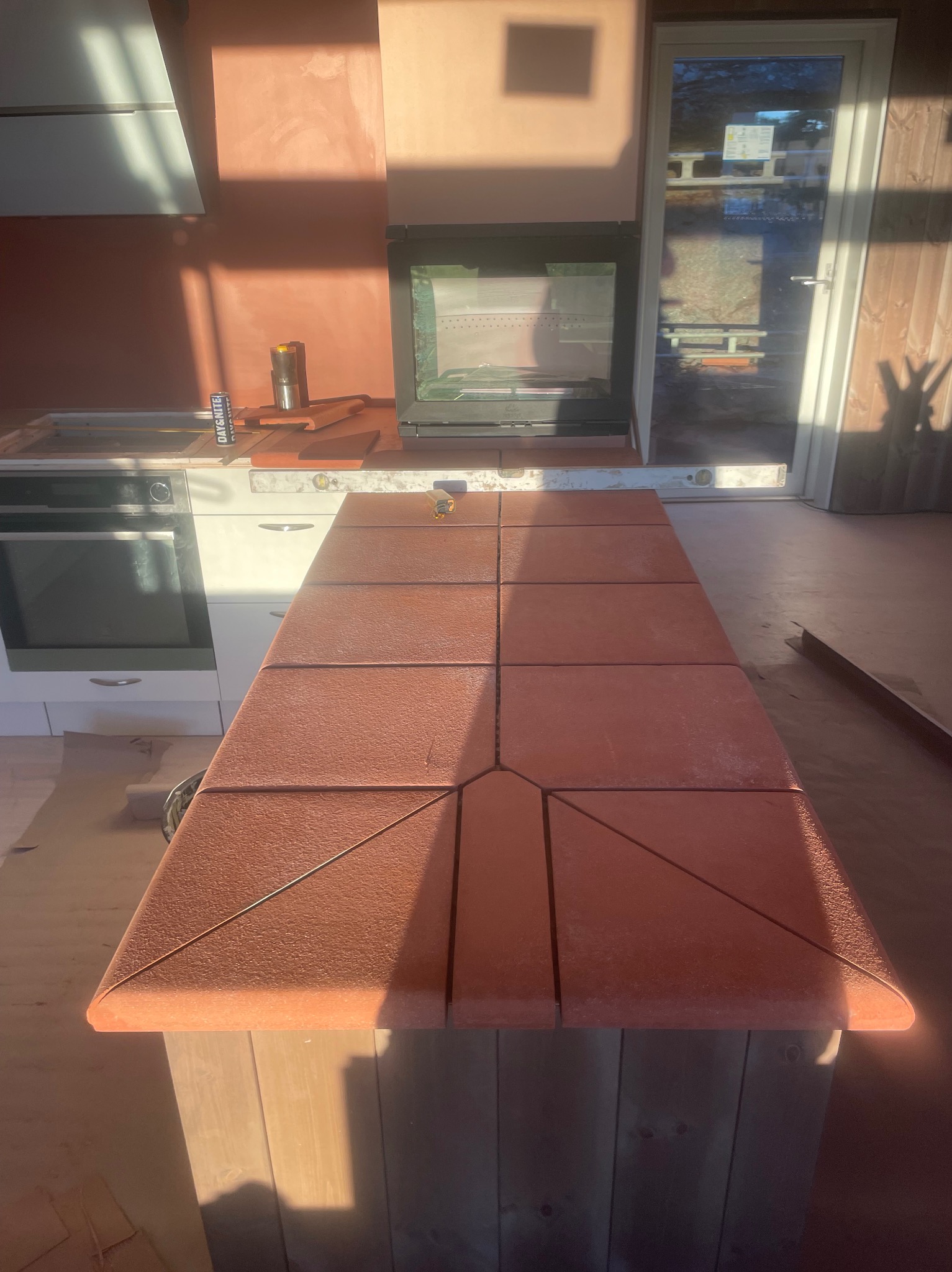 Ceglana wyspa kuchenna w trakcie montażu, z widocznym poziomowaniem. W tle ściana z fakturą i kominek. Jasne światło słoneczne podkreśla detale.