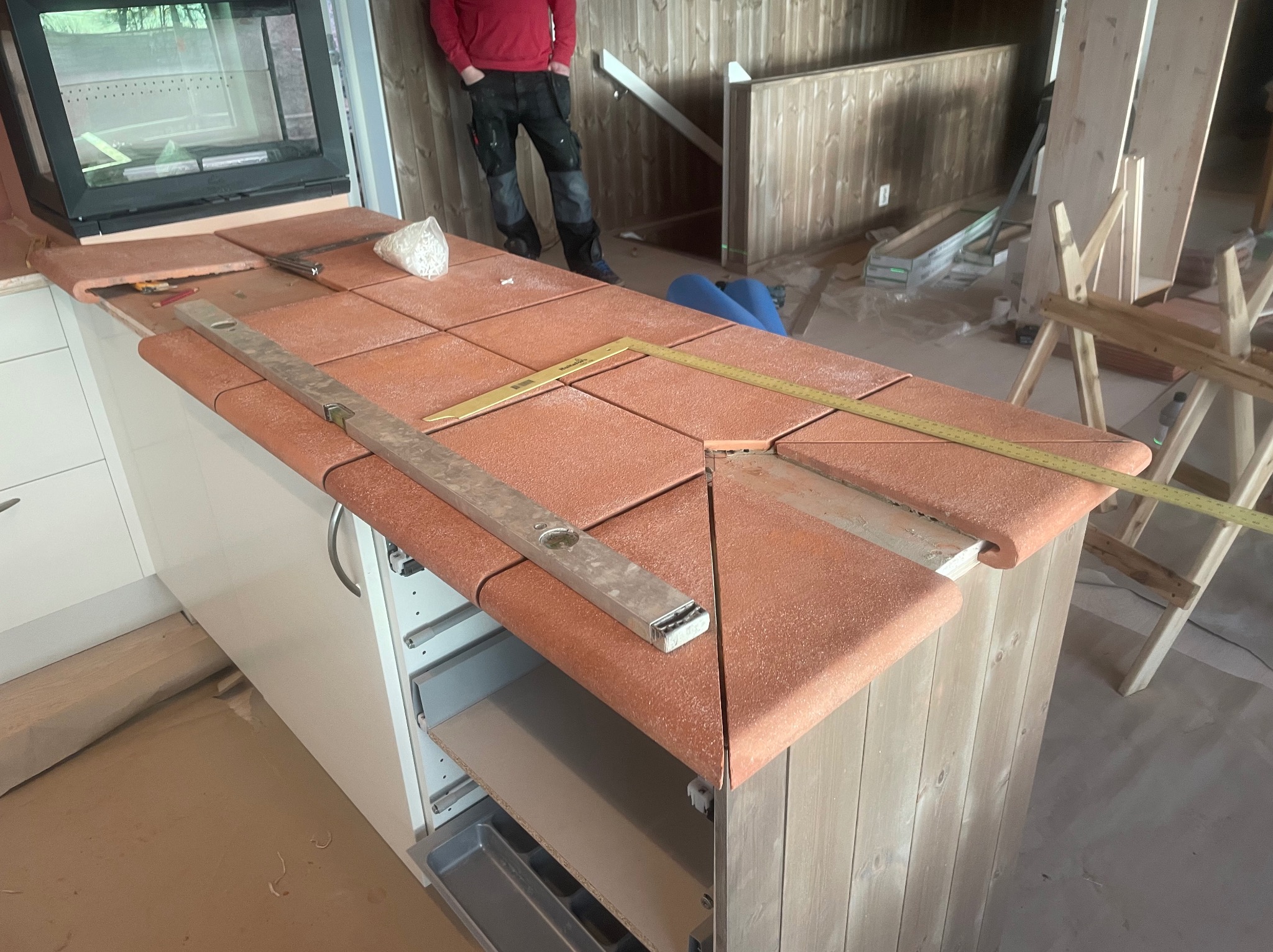 Montaż terakoty na blacie kuchennym. Widoczne poziomica, miarka i białe szafki. W tle fragment pieca i osoba w czerwonej bluzie. Ujęcie z bliska, w trakcie prac.