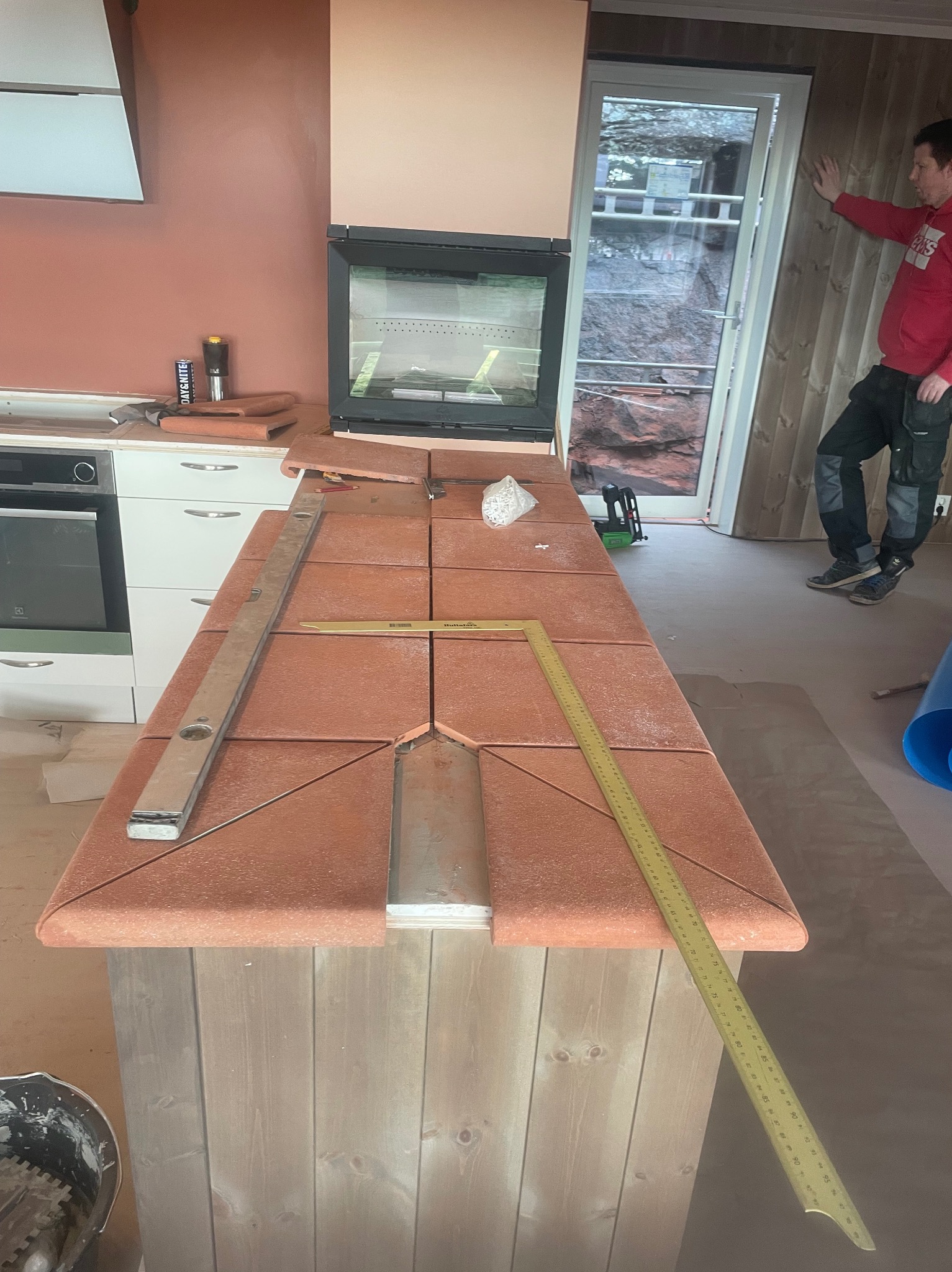 Blat kuchenny z terakoty w trakcie montażu, widoczne narzędzia: poziomica i miarka. W tle okno i osoba w czerwonej bluzie. Nietypowe wykończenie.