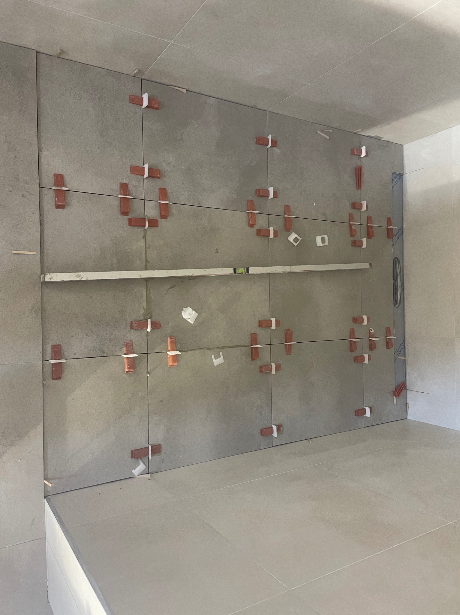 Ściana w trakcie układania szarych płytek, z użyciem poziomicy i krzyżyków dystansowych. Widoczne fragmenty podłogi wyłożonej jasnymi płytkami.