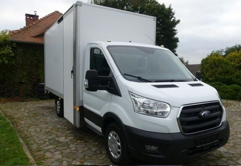 Biały furgon Ford Transit z zabudową kontenerową na brukowanym podjeździe, widok z przodu i boku, zieleń w tle.